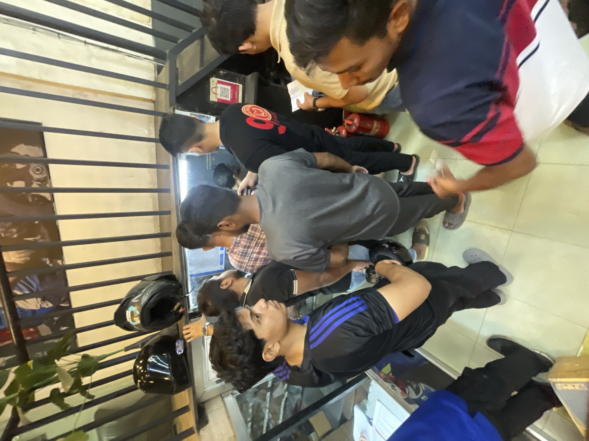 The image shows a group of people inside what appears to be a shop or retail space. The perspective is from an elevated angle, looking down on the group. Several individuals are gathered together, seemingly engaged in a conversation or transaction. Some are standing while others are crouched or leaning.
The environment appears to be enclosed, with a metal grid or railing visible in the background. Helmets are present, suggesting the shop may be related to motorcycles or similar equipment. The interior appears brightly lit.
The location of Dhaka, Bangladesh, is not explicitly indicated within the visible elements of the image. Further information would be needed to confirm this detail.