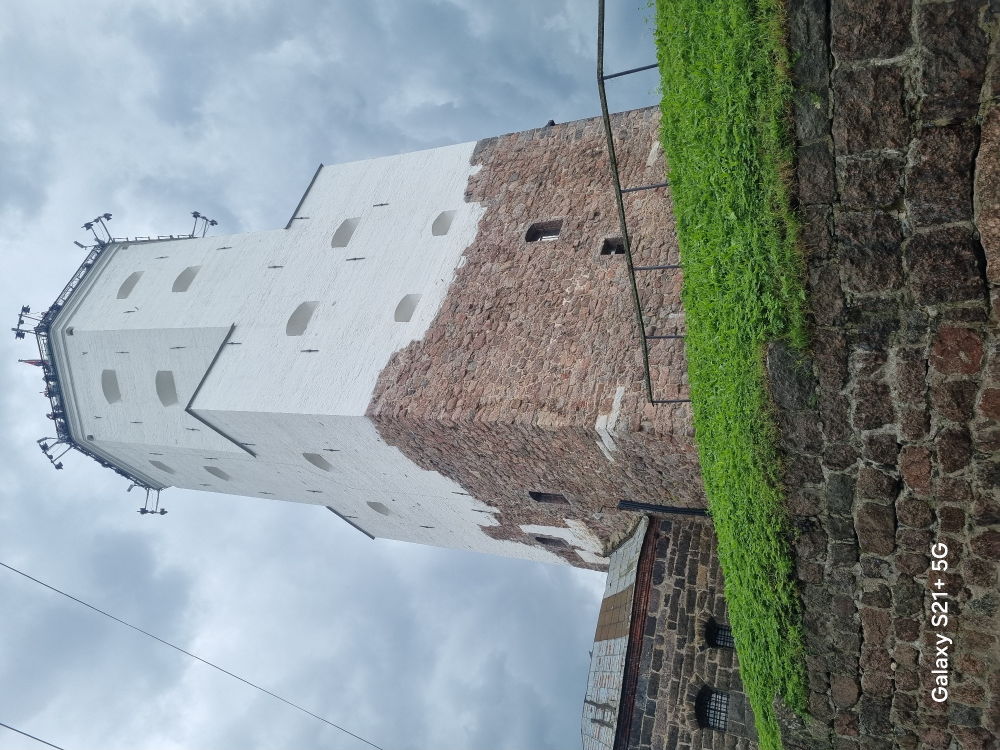 The image depicts a large, white-washed tower with a red stone base, likely part of the Vyborg Castle complex in Vyborg, Russia. The tower has several small, arched windows along its upper section and larger, rough-hewn windows in the stone base. At the very top, there's a black railing with some equipment mounted on it, and a few people are visible standing on the platform. The tower is surrounded by green grass on a slope leading down to a stone wall in the foreground. To the left of the tower, a section of a smaller building with a weathered, metal roof and stone walls is visible. The sky is overcast with dark, cloudy conditions. The bottom of the image shows the text "Galaxy S21+ 5G".
