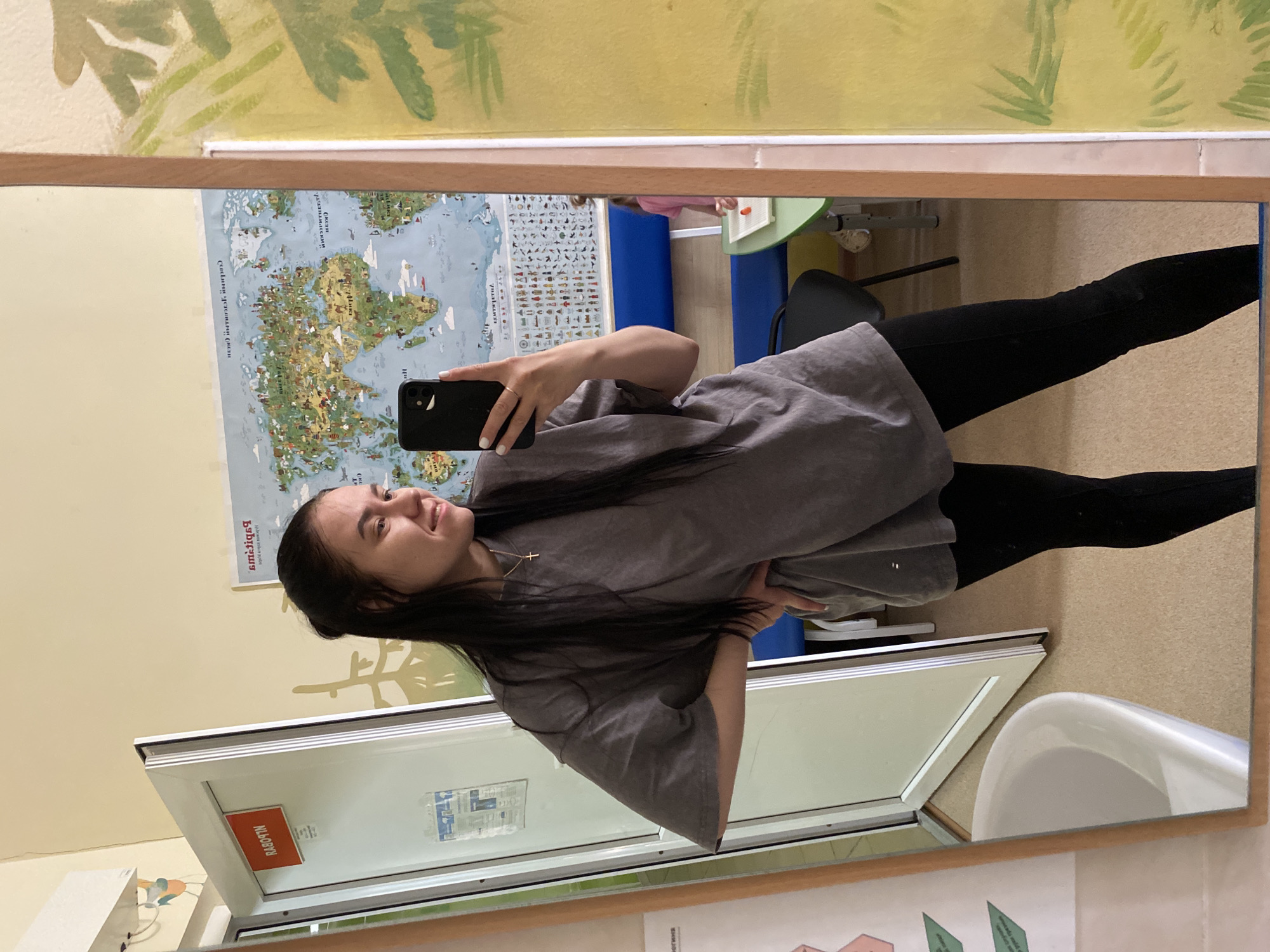 A young woman stands in front of a mirror, taking a selfie. She is wearing black leggings and a grey t-shirt. In the reflection, a world map is visible on the wall, along with a whiteboard and a window. Outside the window, a building is visible. Based on the architectural style and presence of Cyrillic writing, the location is likely Perm, Russia. A sink is also visible in the reflection, suggesting a bathroom or classroom setting.