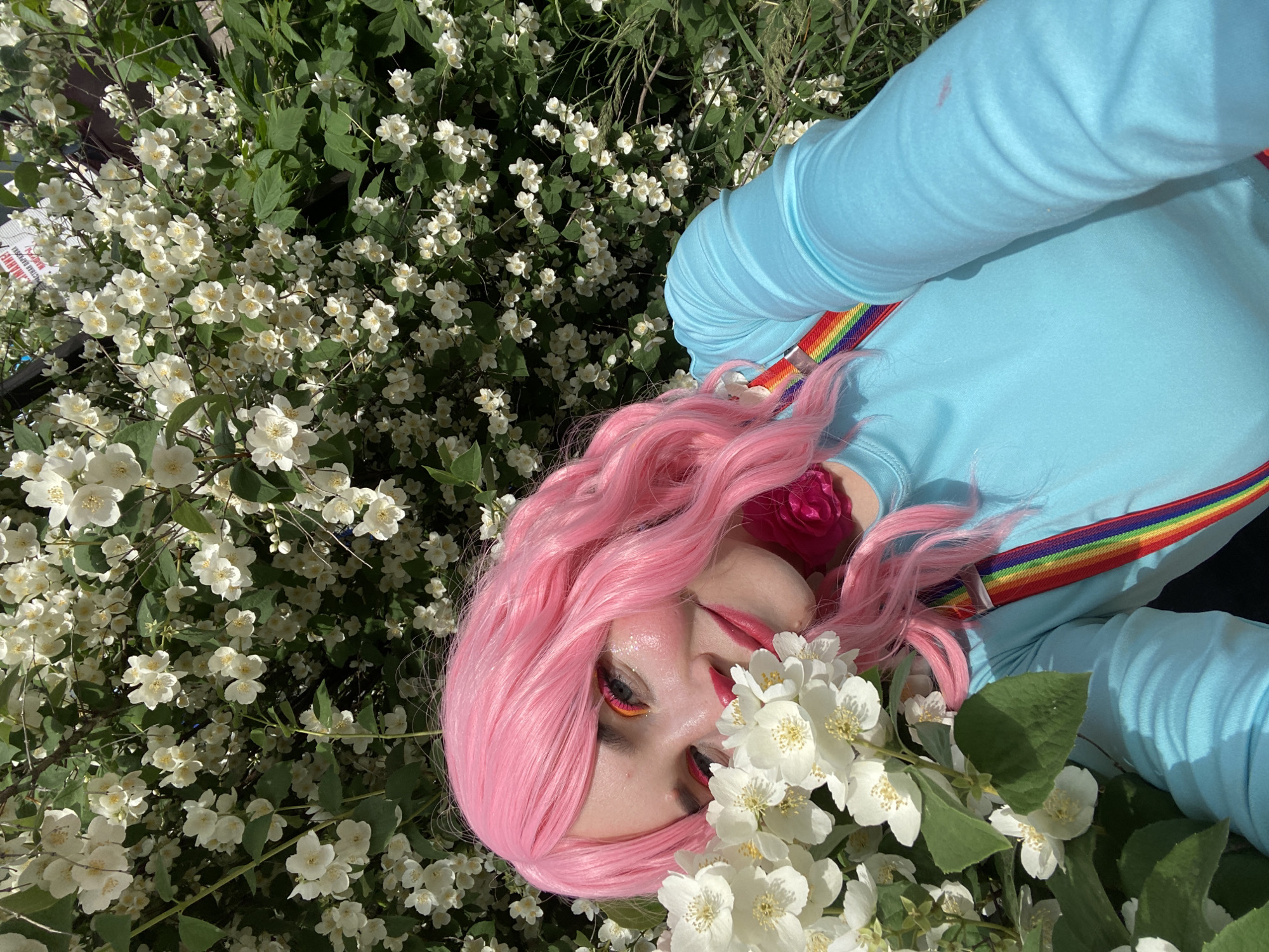 A young woman with bright pink, wavy hair and dramatic eye makeup is taking a selfie, partially obscured by a bush of white jasmine flowers. She is wearing a light blue long-sleeved shirt with rainbow suspenders. Her makeup includes pink blush, pink lipstick, and brightly colored eyeshadow in pink and orange tones. A dark pink accessory, possibly a collar or bow, is visible around her neck. The background is dominated by lush green foliage and the white jasmine blossoms, suggesting an outdoor setting in a park or garden. A blurry sign with Russian text is visible in the upper right portion of the image. The scene appears to be set in Chaikovsky, Russia, as indicated by the provided context.