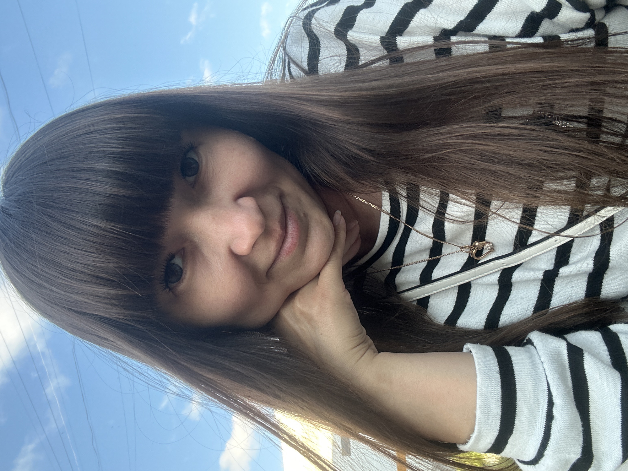 A young woman with long, dark hair and bangs is pictured in an outdoor, urban setting, possibly in Chaikovsky, Russia. She is wearing a black and white striped sweater and a delicate necklace with a heart-shaped pendant. Her hand is resting on her chin, and she is looking towards the camera with a gentle smile. The background shows a bright blue sky with some wispy clouds and power lines, suggesting an open area within a city.