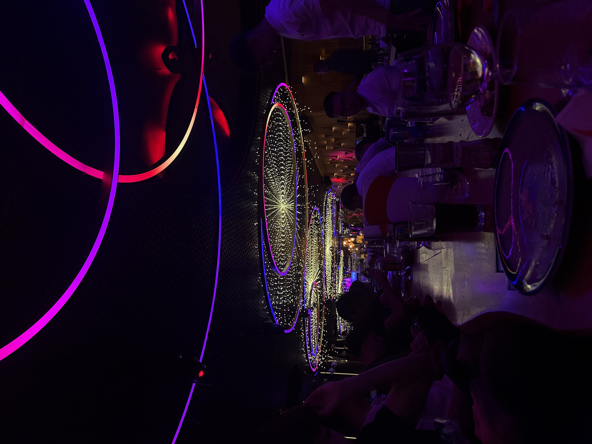 The image depicts the interior of a dimly lit, modern restaurant or club in Sofia, Bulgaria, characterized by vibrant neon lighting. The ceiling features elaborate circular light installations with thousands of small white lights, accented by rings of purple and blue neon. A long wooden table is set with plates, glasses, and what appear to be drinks. Several patrons are seated at the table, mostly in silhouette or partially obscured by the low light and their attire. Some individuals are looking down at the table, possibly eating or socializing, while others are standing or walking in the background. The overall ambiance suggests a lively evening setting with a focus on contemporary design and atmospheric lighting.