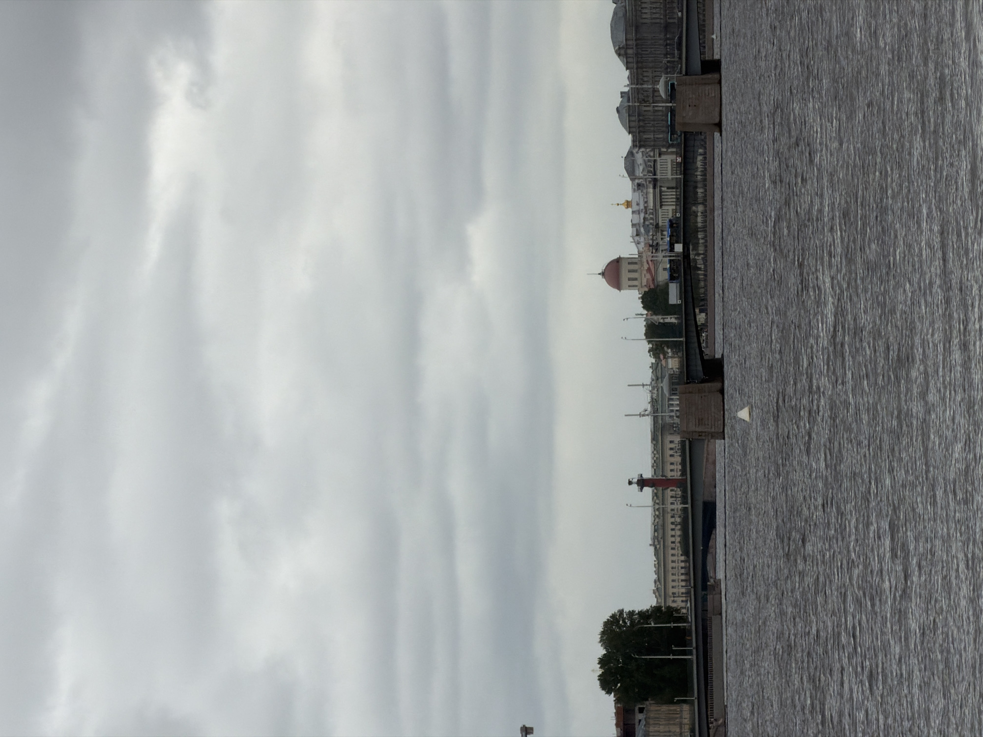This is a landscape photograph of the Neva River in Saint Petersburg, Russia, under a heavily clouded sky. The river occupies the foreground, with choppy, dark water reflecting the overcast conditions. In the middle ground, a bridge spans the river, with its stone abutments visible. Beyond the bridge, the cityscape of Saint Petersburg is seen. Prominent buildings include a structure with a red dome and a golden dome, along with other classical architectural styles. A tall, red rostral column, a landmark of Saint Petersburg, stands to the left of the dome buildings. Trees and streetlights are also visible along the embankment. The overall impression is a cool, subdued atmosphere typical of a cloudy day in Saint Petersburg.