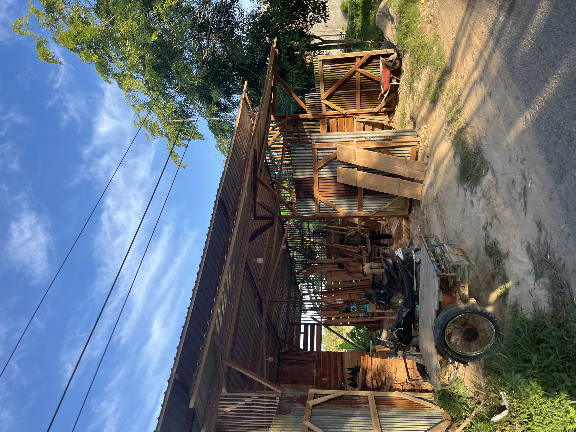 The image depicts an outdoor scene at what appears to be a woodworking area or workshop in Seuneubokpunti, Indonesia. The setting is rustic, with a partially constructed or open-air wooden structure covered by a corrugated metal roof. Timber is stacked in various locations, suggesting ongoing construction or woodworking activities.
Several objects are visible, including a three-wheeled utility vehicle, possibly used for transport of materials. There's also a red wheelbarrow near the wooden structure. The ground is a mix of dirt and some patches of grass. In the background, trees with green foliage are present, indicating a somewhat rural or natural environment. The sky is blue with wispy white clouds, suggesting a clear day, likely in the late afternoon or early morning given the quality of light and shadows.
Two individuals are visible within the workshop structure. One person, partially obscured, appears to be working with wood. Another person, shirtless, is also in the vicinity, possibly involved in the same activity.
Notable details include the presence of power lines running overhead, a common sight in many parts of Indonesia. The overall atmosphere seems industrious and practical, with the focus on the woodworking operations. There is no visible text in the image.