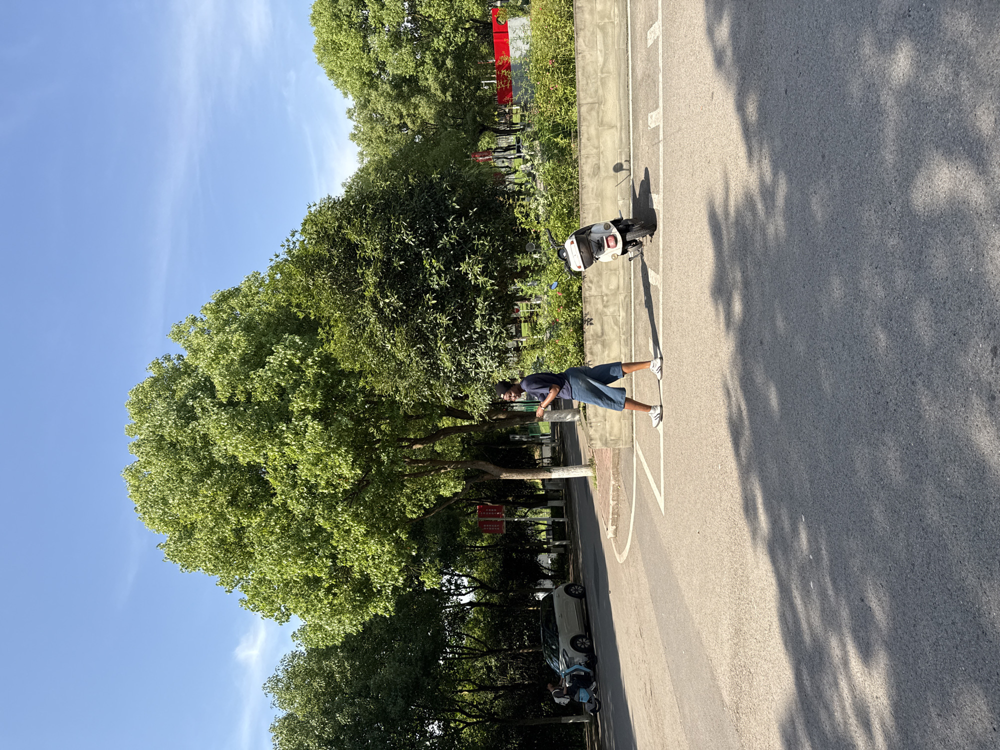The image captures a sunlit outdoor scene, oriented as if viewed along a tree-lined paved path or road. The sky above is a clear, vibrant blue with a few faint, wispy clouds. Strong, dappled shadows from the trees stretch across the asphalt, indicating bright sunshine and suggesting it is daytime, likely late morning or afternoon.
On the right side of the paved surface, a man stands, dressed in a dark blue short-sleeved top, blue shorts, and white sneakers. He appears to be looking towards the left side of the frame. Further to his right, parked on a concrete curb, is a white scooter or moped. The lower legs and white sneakers of another person are partially visible next to the scooter, hinting at a second individual.
The left side of the path is densely bordered by tall, leafy green trees that provide extensive shade. Smaller green bushes also line the right-hand curb. In the midground, nestled among the trees on the left, a light-colored car is partially visible. A red sign with white Chinese characters is mounted on a pole amidst the greenery on the left, but the text is not clearly legible. The asphalt surface of the road is marked with distinct white lines, including solid borders, dashed segments, and a partial circular marking.
The scene evokes a peaceful, everyday atmosphere, set in what appears to be a park, campus, or quiet street in Jiangjin, China.