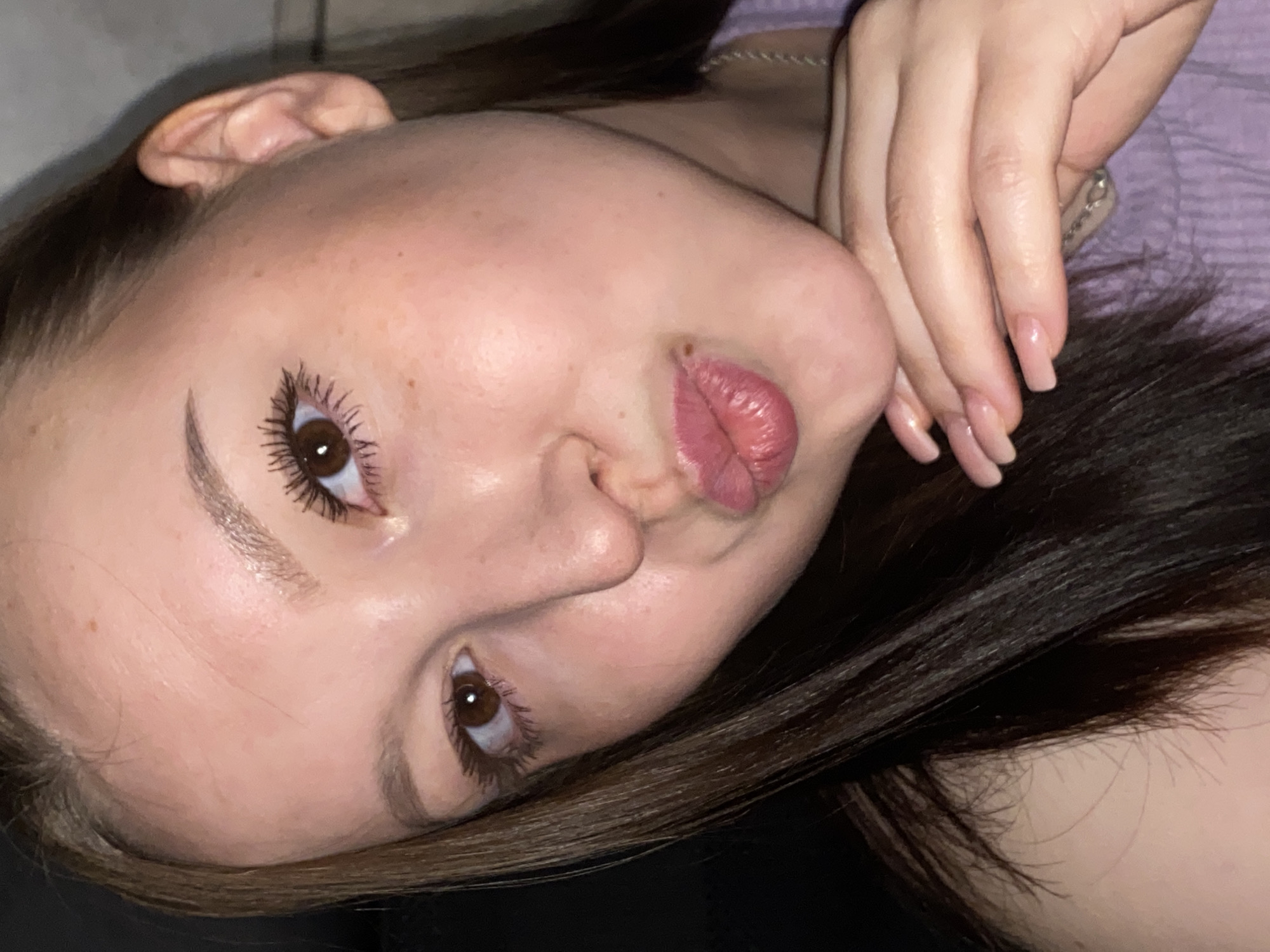 The close-up, high-angle shot captures the upper body and face of a young woman, lying down or reclining, with her head tilted. Her face is the primary focus, showcasing her brown eyes, defined eyebrows, a small nose, and puckered lips with pink lipstick. Mascara is visible on her long eyelashes. Her skin appears clear with a few small freckles on her nose and cheek.
Her long, dark brown hair frames her face and extends below her. One hand is resting near her cheek, revealing long, well-kept fingernails with a natural or light nude polish. She is wearing what appears to be a light purple, possibly ribbed, top or blanket, and a thin silver-colored chain necklace is visible around her neck.
The background is simple and out of focus; dark beneath her head and a lighter, indistinct surface above. The lighting suggests an indoor setting. The overall expression conveyed by her puckered lips and direct gaze is playful or mischievous. There is no visible text in the image.