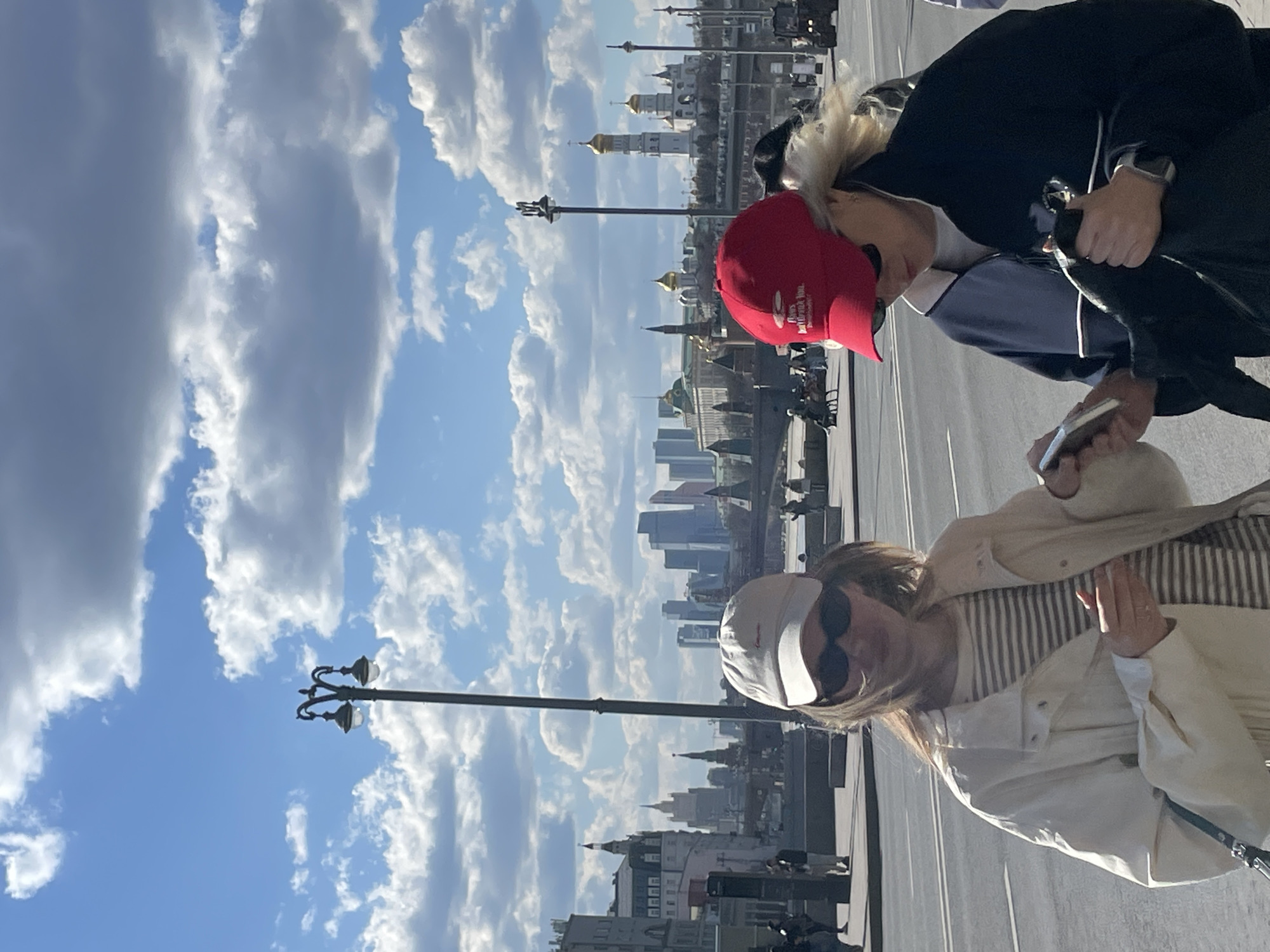 Two women are captured in a photograph against a backdrop of Moscow's city skyline and a cloudy sky. One woman is wearing a white jacket, sunglasses, and a cap, while the other is wearing a red cap, a dark jacket, and holding a phone. Both individuals appear to be in an urban setting, possibly a sidewalk or pedestrian area, with buildings and lampposts in the background. The city skyline, featuring Moscow's distinct architecture, including golden domes, is partially visible. The sky is filled with white clouds, enhancing the overall outdoor setting.