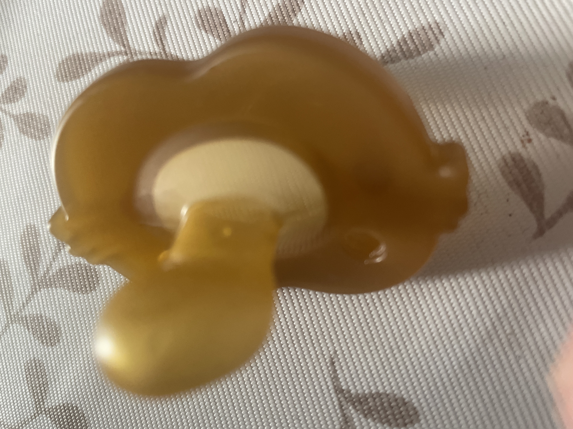 A close-up, slightly out-of-focus, overhead shot displays a single, amber-colored pacifier on a textured, white and brown patterned surface. The pacifier is positioned on its side, with the nipple extending to the left. The surface appears to be a placemat or tablecloth with a floral design. The image does not provide any information or context related to the city of Otradnoye, Russia, or any other specific geographical location.