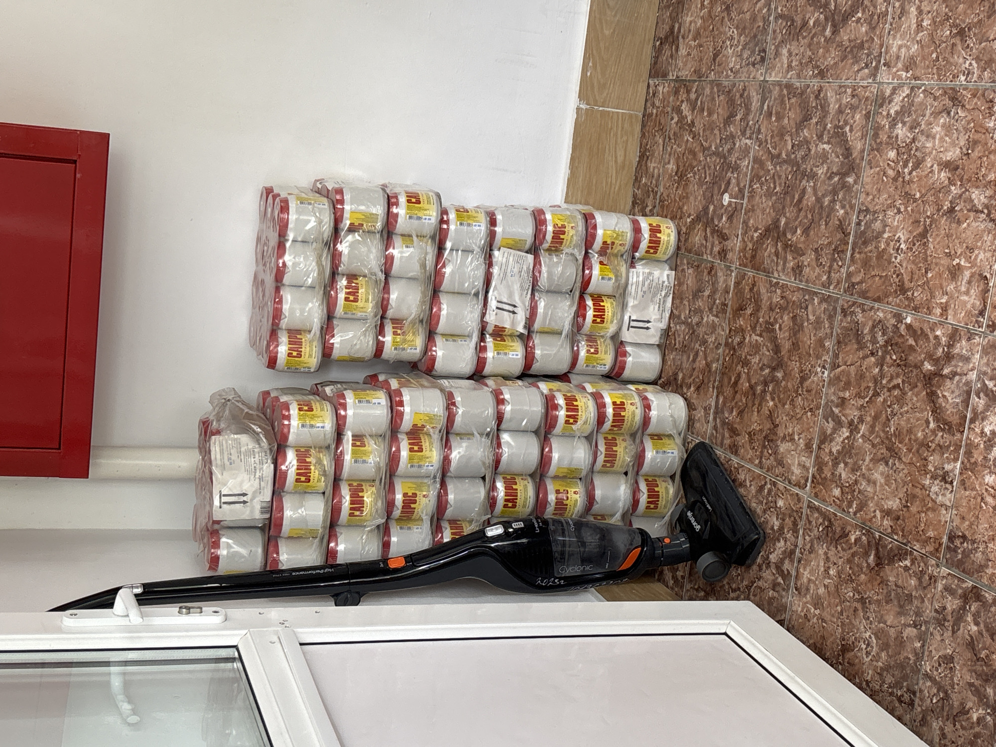 In the city of Unknown, Unknown, an indoor scene is depicted, likely a storage area or a hallway, with a focus on a stack of canned goods and a vacuum cleaner. The floor is covered with brown marble-patterned tiles. To the left, a white door with a white handle is partially visible. A black upright vacuum cleaner, identified by the "Gorenje" logo and the text "High-Performance Cordless," is positioned vertically next to the door.
Dominating the right side of the image is a tall stack of canned goods, shrink-wrapped in clear plastic. The cans are white with red lids and yellow labels. The labels feature red lettering that appears to spell "CANPOC" and also includes some Cyrillic text. Some of the top cans have shipping labels with black markings and arrows. The stack reaches a significant height, suggesting a substantial quantity of product.
In the background, a white wall extends upwards, and a portion of a red structure, possibly a cabinet or a fire extinguisher box, is visible at the top of the frame. The lighting in the scene appears to be artificial, casting subtle shadows. There are no people present in the image.