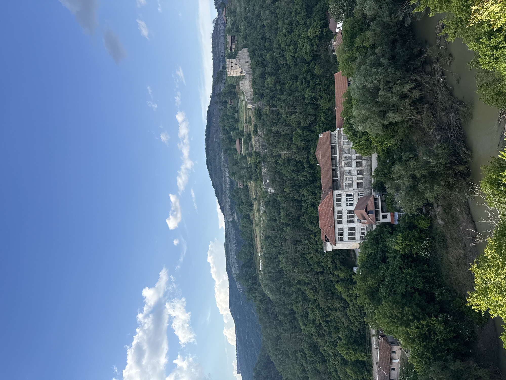 The image depicts a landscape in or near Veliko Tarnovo, Bulgaria. A large, tree-covered cliff face dominates the scene, rising sharply on the left side of the frame. A multi-story building with a red-tiled roof is nestled among the trees on the right side. The Yantra River flows on the right edge. The sky, blue with scattered clouds, occupies the upper left portion of the image. Remnants of an ancient stone structure are visible higher up on the cliff.
