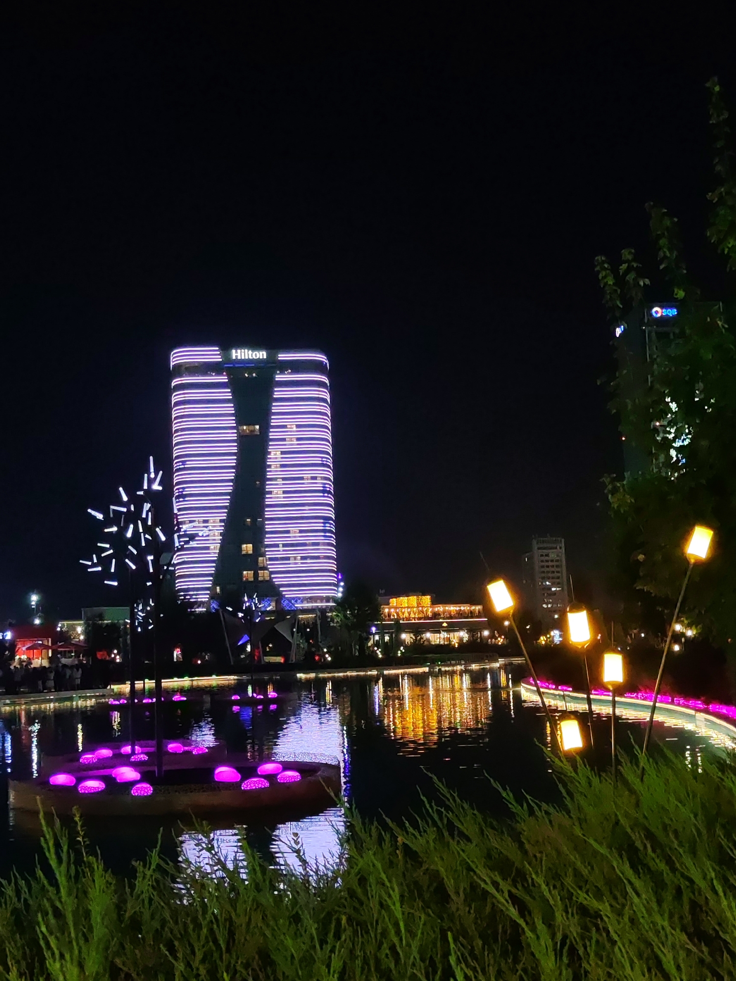The image depicts a nighttime scene in Tashkent, Uzbekistan, likely at the Magic City entertainment complex. A prominent Hilton hotel, illuminated with horizontal purple lights, dominates the background. To the left of the hotel, a unique art installation resembling a stylized tree with numerous white lights is visible. In the foreground and midground, a body of water reflects the city lights. Several decorative lamps with yellow, square shades are spaced out along the edge of the water. On a small island in the water, there are glowing purple objects, possibly decorative lights or seating. The overall ambiance is one of vibrant urban nightlife, with a focus on illuminated architecture and decorative elements.