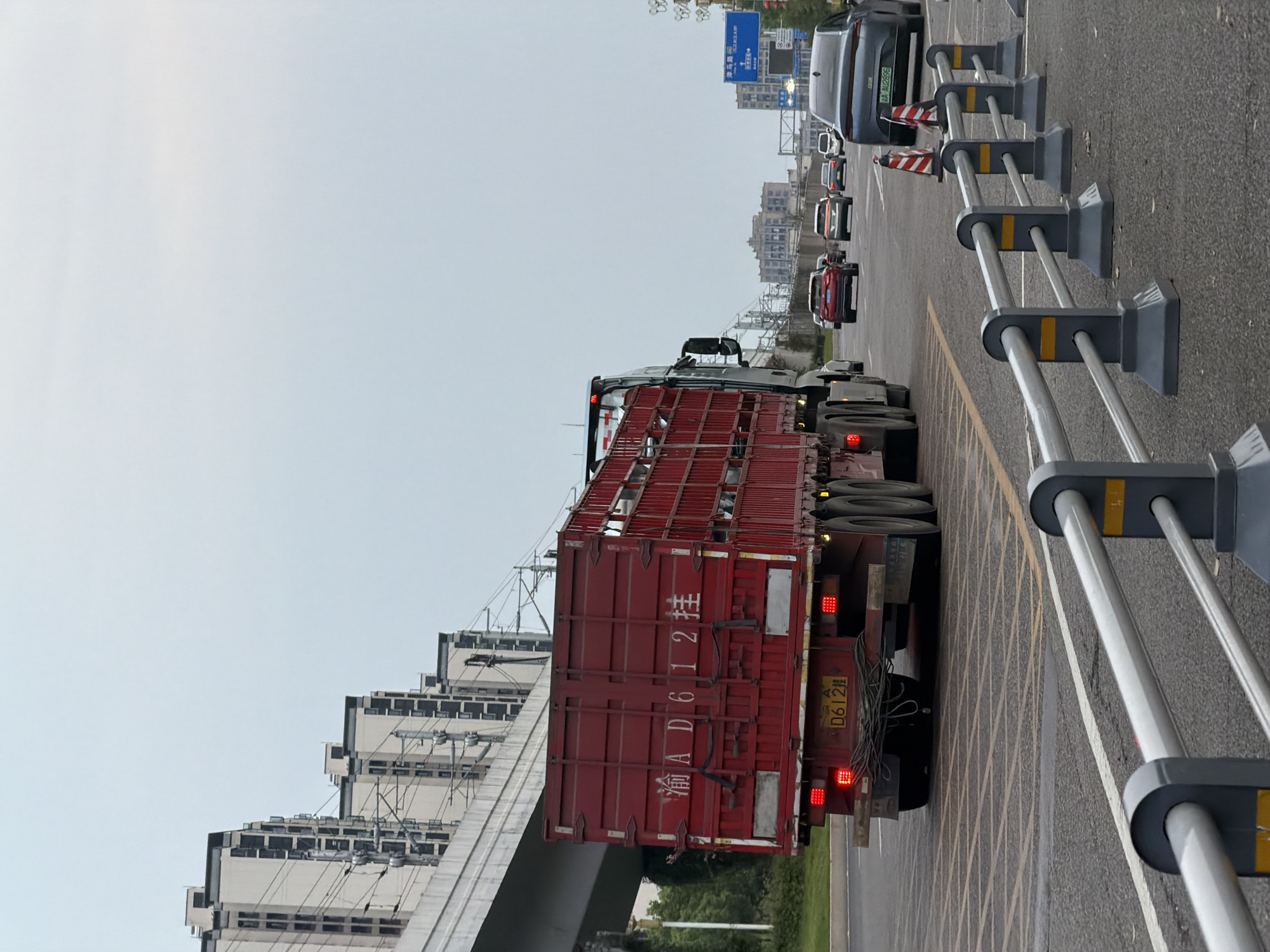 An overcast, gray day frames a street scene in Jiangjin, China. A large red truck, with the license plate "渝 AD612 桂", dominates the foreground, traveling on a multi-lane road. The truck's cargo container is open at the top, revealing a framework of metal bars. Behind the truck, several cars are visible in traffic, including a dark gray sedan with a license plate that reads "A·MZ695". The road is marked with yellow and white lines, and temporary traffic barriers line the right side of the image. In the background, a modern elevated train track runs parallel to the road, with rows of tall, apartment buildings visible beyond it. Further in the distance, more buildings and the hazy outlines of other structures can be seen. Traffic signs are present on the right side of the road.