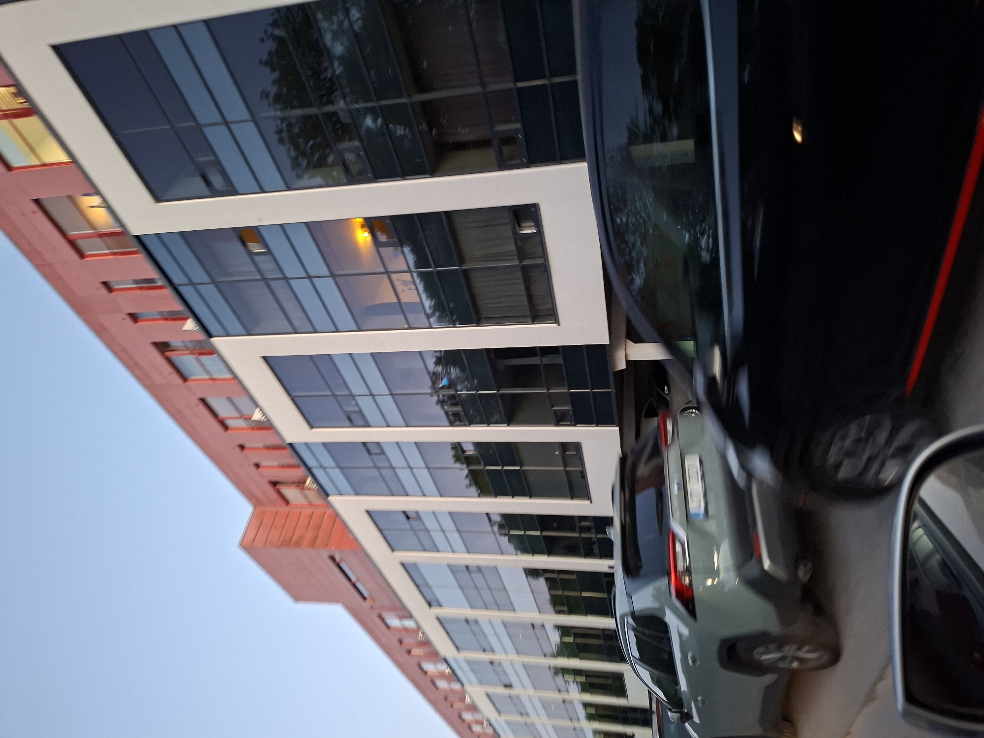 The image depicts a street scene in Tallinn, Estonia, specifically along Akadeemia tee in the Tehnopol Science Park. The primary subject is a multi-story building characterized by a distinct red/terracotta upper section with smaller, recessed windows and a modern, vertically extended lower facade composed of large, dark-paned glass windows framed by white structural elements. Reflections of trees and a clear, light blue sky are visible in the glass. Several windows in the lower section display an internal warm yellow illumination.
In the immediate foreground, a dark-colored SUV or car, likely black, is captured in motion blur, presenting a partial view of its side and a distinct red stripe running along its lower body. To the left, in the midground, a light green/grey Toyota RAV4 SUV is visible from the rear, also exhibiting some motion blur. Its Estonian license plate, "477 TLL," is partially legible. A silver/grey vehicle is partially visible behind the RAV4. The road surface appears to be asphalt. The scene suggests an urban setting during daylight hours, possibly late afternoon or early evening, given the sky's color and internal lights. No individuals are visible in the image.