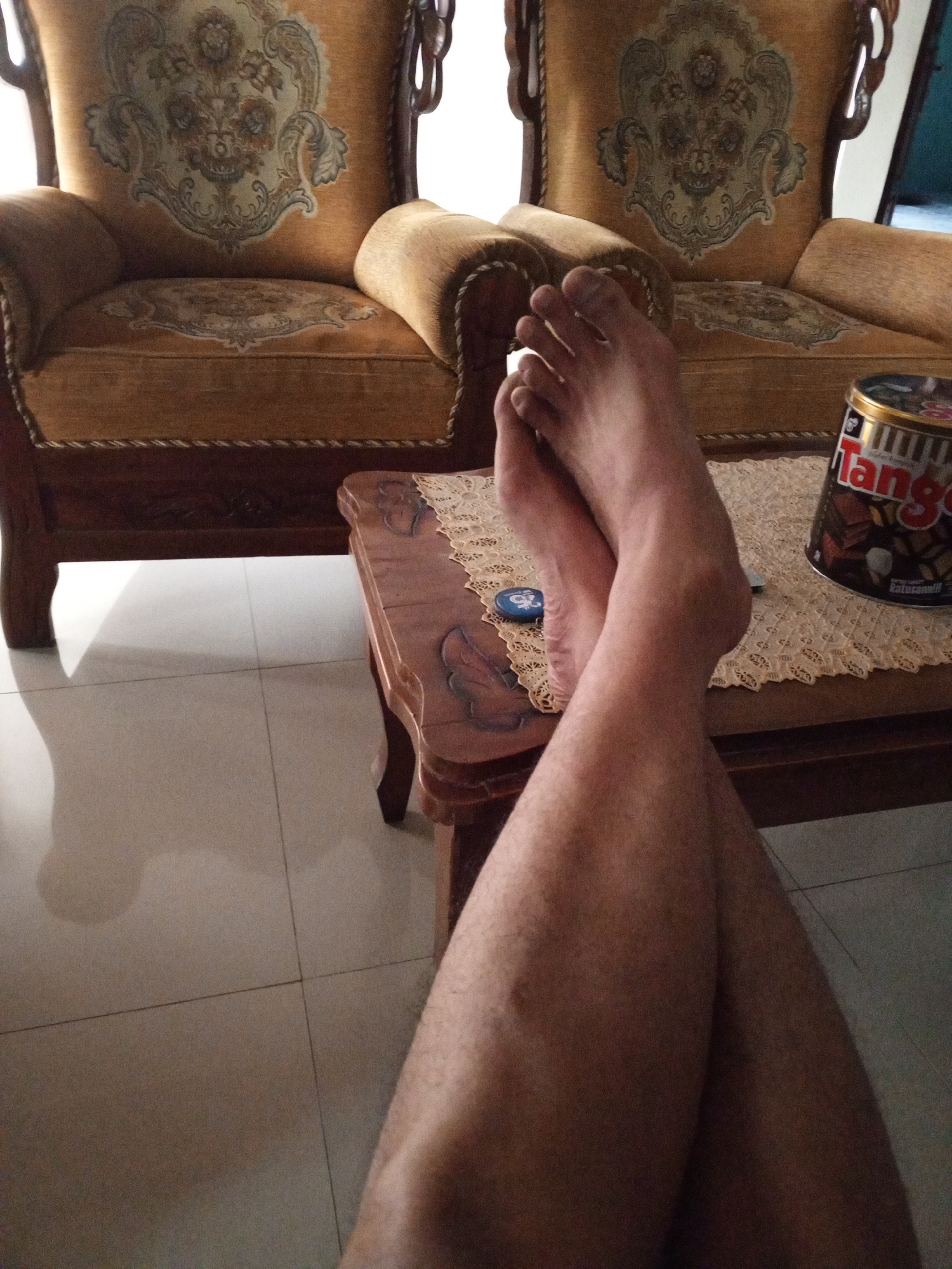 The image depicts a still life with a pair of crossed legs resting on a coffee table. The legs, belonging to an unseen person, are positioned in the foreground, with the feet visible and oriented towards the left. The individual's skin tone is light to medium.

The coffee table is made of dark wood and has an intricately carved edge. On its surface, a beige lace doily is spread, and on top of that sits a cylindrical metal tin. The tin, labeled "Tango" with text in Indonesian, contains a dark-colored product, likely snacks or biscuits, with visible images of chocolate and wafers. A small blue object, possibly a cap or a button, is also on the doily near the feet.

In the background, two ornate armchairs are visible. They are upholstered in a golden-brown fabric with detailed floral patterns. The armchairs are substantial, suggesting they are part of a living room or lounge area. The floor tiles are large and off-white, reflecting the ambient light. Shadows are cast on the floor, indicating a light source from the right.

The scene is indoors, likely a residential setting in Cabeyan, Indonesia, given the context provided. The overall impression is one of relaxation and casual enjoyment, possibly at home. There are no other people or animals present in the frame.