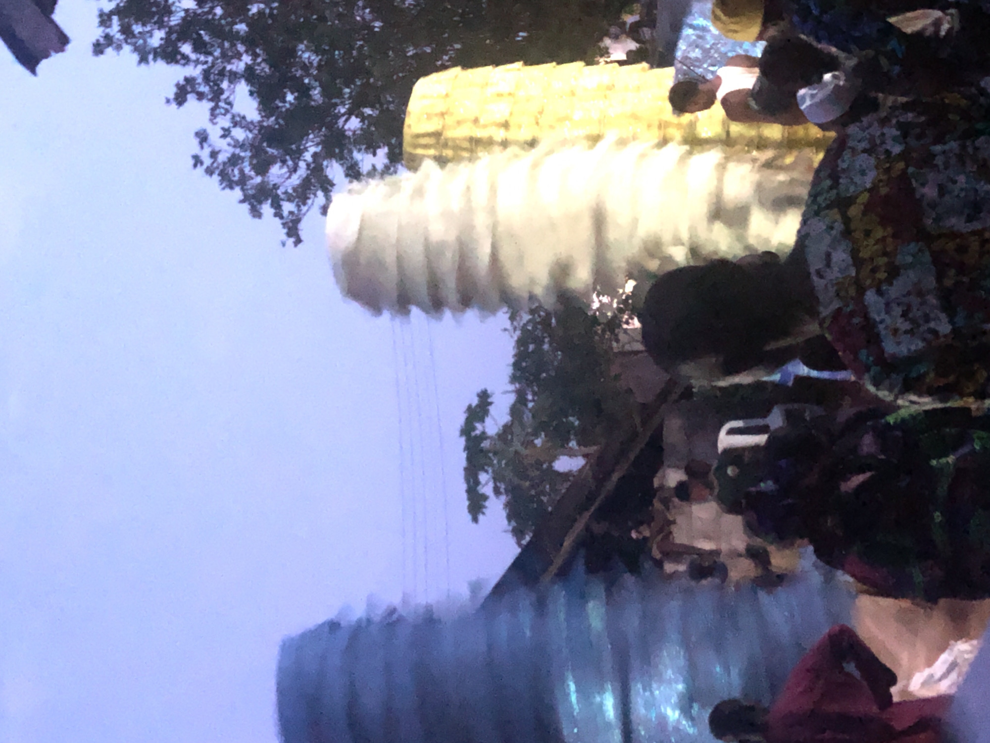 The image depicts an outdoor scene in Unknown, Unknown, featuring a crowd of people gathered near trees and buildings. At the top of the image are trees silhouetted against a light blue sky. Two large, cylindrical, stacked structures dominate the image; one is yellow and the other white. The bottom of the image is dominated by a blue structure of stacked cylinders. People are positioned in the foreground, with some partially obscured or out of focus. The people appear to be observing the stacked cylinders.