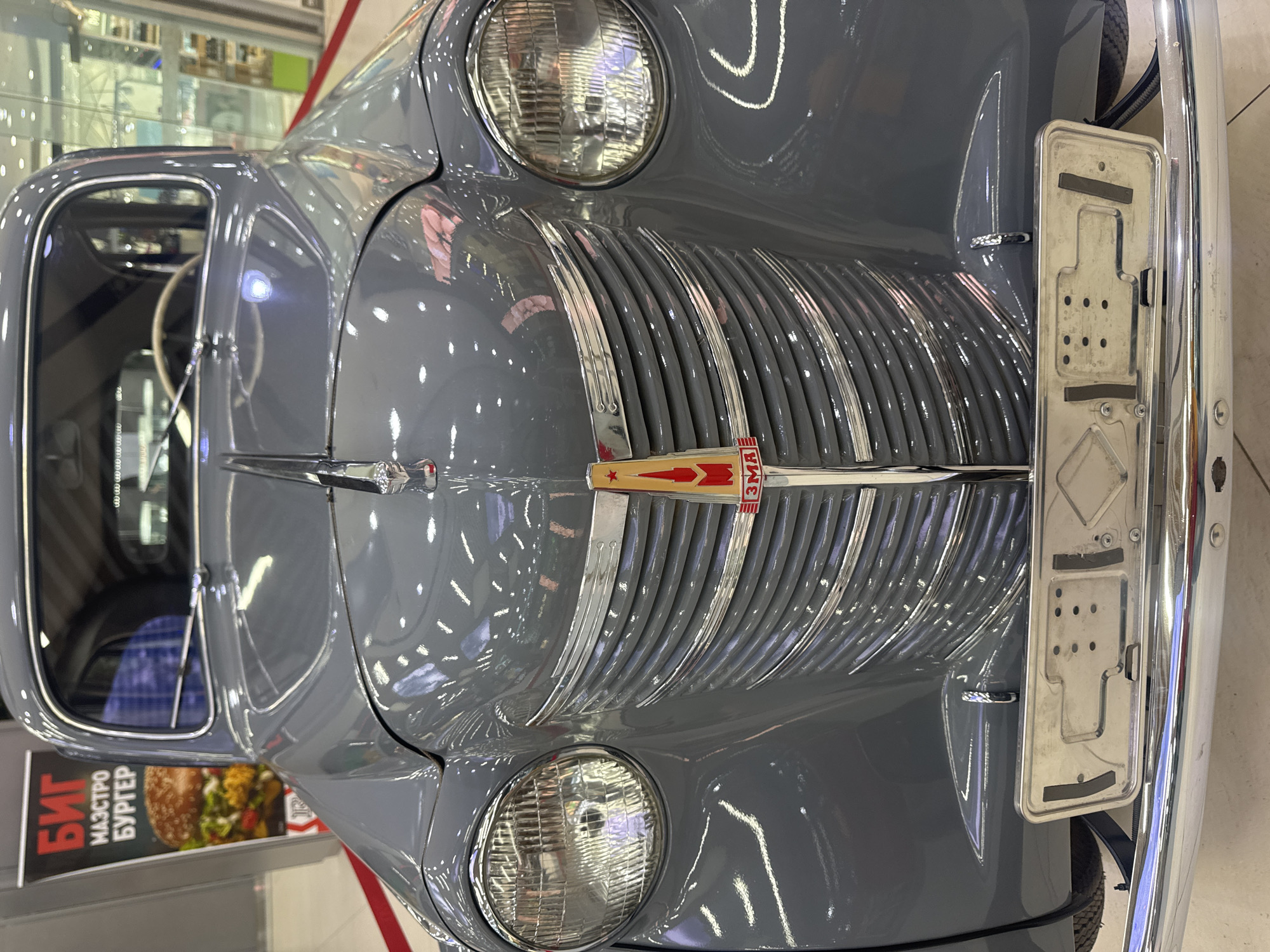 A close-up, eye-level, frontal view of a vintage grey car is captured. The vehicle's chrome grille, headlights, and hood ornament are prominent. The hood ornament features a red and yellow emblem with a star. The car's license plate holder is visible at the bottom. In the background, a banner with Russian text and an image of a burger is partially visible. The setting appears to be indoors, possibly a mall or exhibition space, indicated by the polished floor, overhead lighting, and reflective surfaces. There are no people visible in the image. The car is parked on a red carpet runner. There is no information in the image to place it in Samara, Russia.