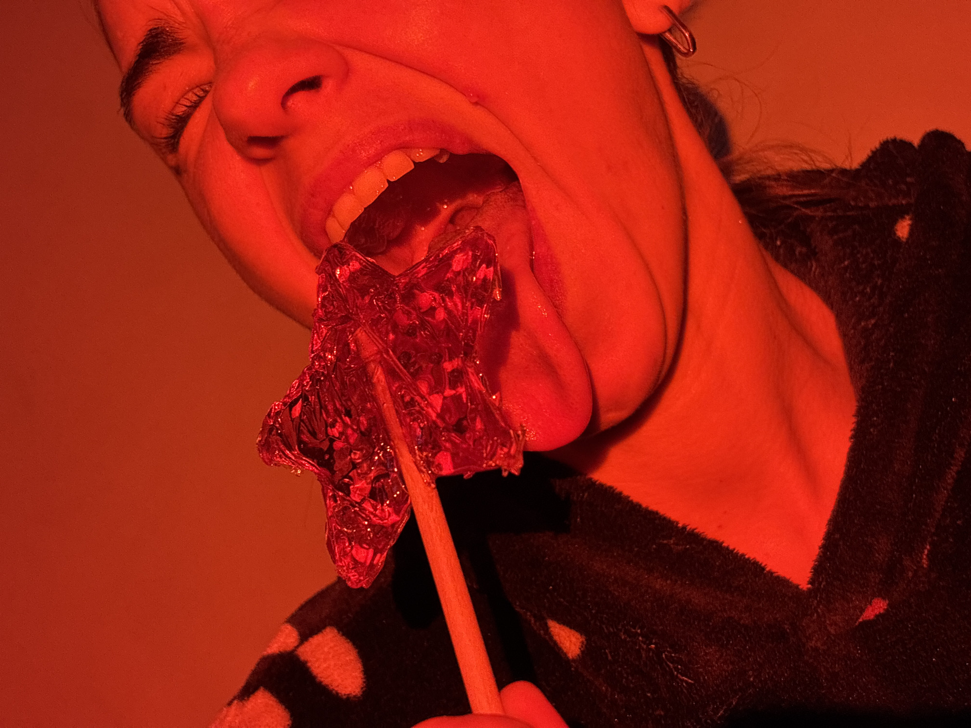 The image shows a person with their head tilted upwards, mouth open wide, and tongue extended, licking a red star-shaped lollipop. The person has fair skin and dark hair, and is wearing a dark, patterned robe or jacket. The background is a solid, uniform red color. The lighting is predominantly red, casting strong shadows and affecting the color perception of all elements in the image. The location is unspecified.
