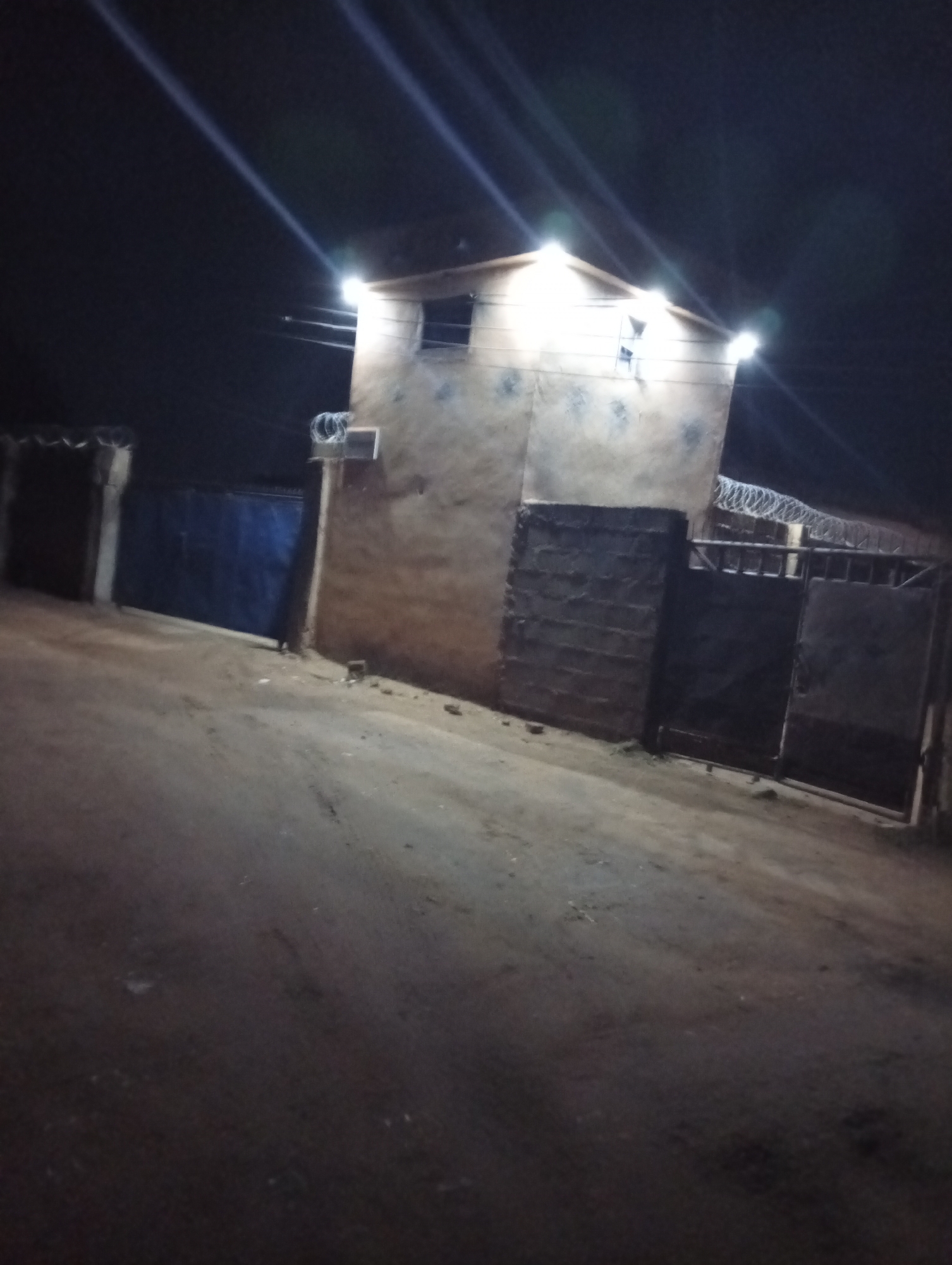 The image depicts a building at night in Jos, Nigeria. The building is rectangular, likely constructed from concrete, and has a flat roof. It is illuminated by several lights. A fence with barbed wire surrounds the property. There is a gate at the entrance. The surrounding area appears to be a dirt road or unpaved surface. The overall environment suggests a low-income or developing area.
