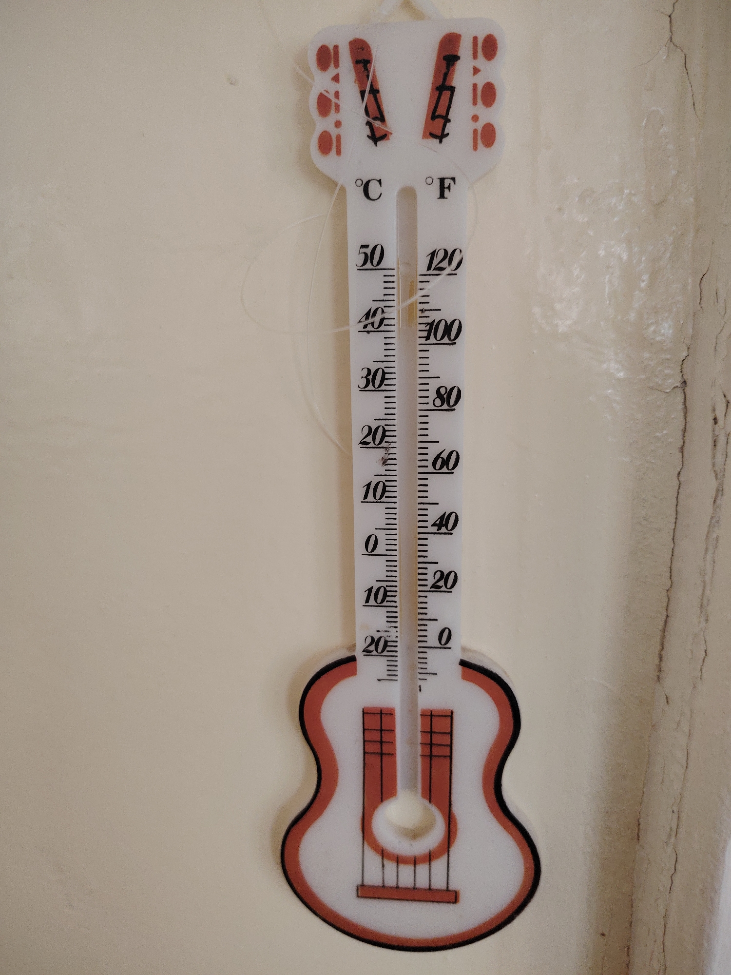 A vertically oriented close-up shot features a guitar-shaped thermometer mounted on a light-colored wall, showing the temperature at approximately 40 degrees Celsius or 100 degrees Fahrenheit. The thermometer, designed to resemble a guitar, has a white body with red accents and a black outline. The temperature scale is positioned in the center of the guitar’s neck, with markings for both Celsius and Fahrenheit readings. To the right of the thermometer, the wall shows some signs of aging, with visible cracks and imperfections. The exact location in Sarapul, Russia is not discernible from the image.