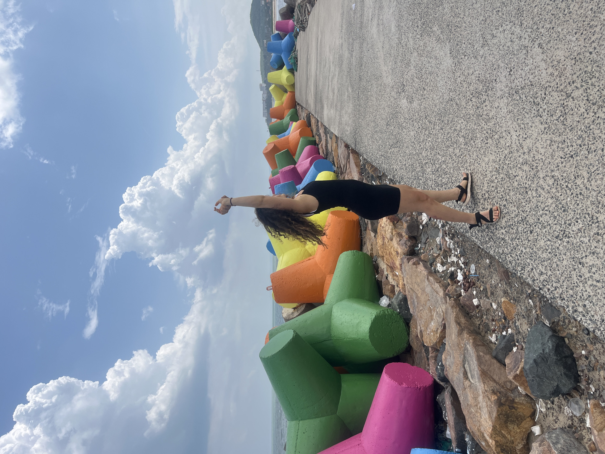 In the city of Phú Thủy, Vietnam, a woman with curly dark hair stands on a paved walkway next to a breakwater made of colorful tetrapods. The woman is wearing a black tank dress and sandals. Her arms are raised above her head, and she appears to be stretching or posing. The tetrapods, a type of concrete armor unit, are painted in bright colors including pink, green, orange, yellow, and blue. They are arranged along the coastline, protecting it from the sea. The sea is visible in the background, with a hazy shoreline and buildings in the distance. The sky is partly cloudy with large, white cumulus clouds against a blue sky. The ground on the left side of the walkway is rocky and covered with debris.