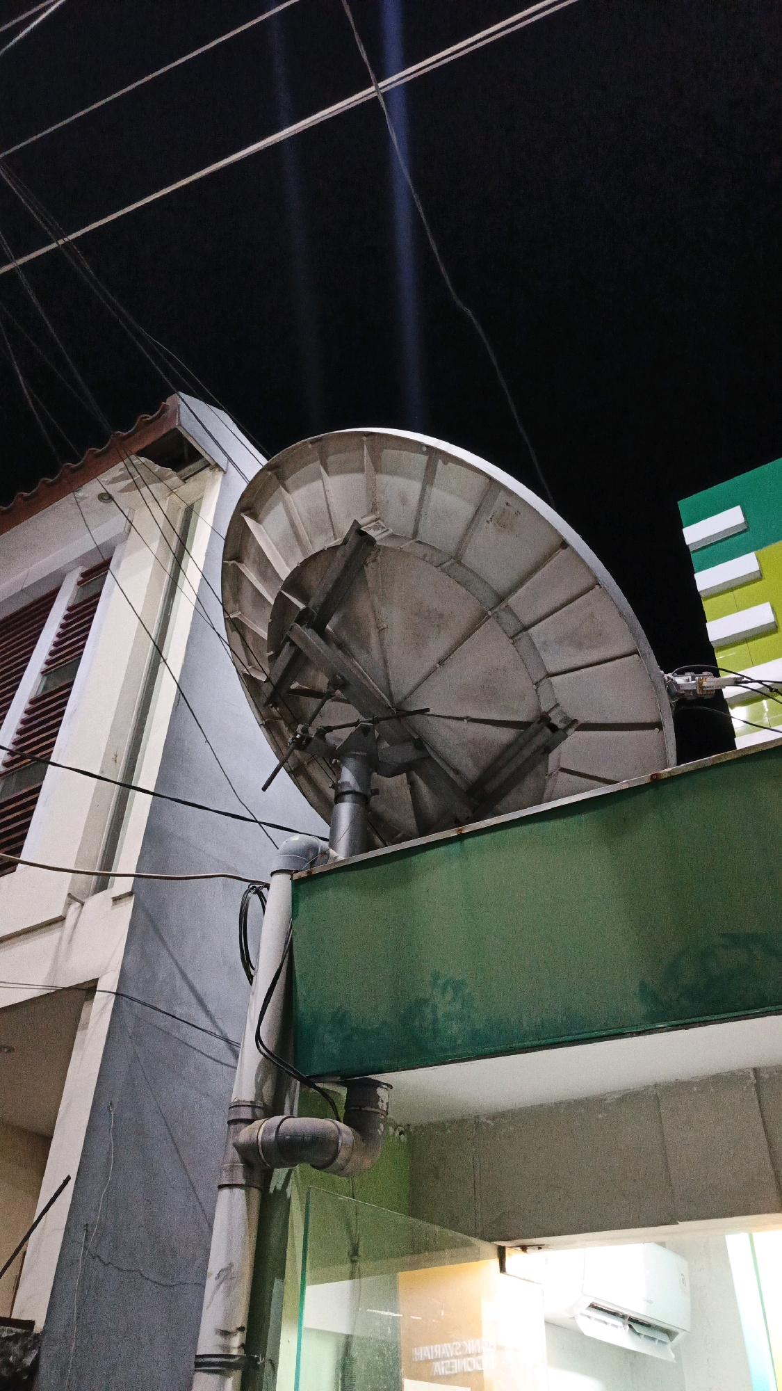 A satellite dish is mounted on the exterior of a building at night in Gondangrejo, Indonesia. The dish is silver and appears to be old and weathered, with a circular reflector and supporting arms. It is positioned on a green ledge that extends from the building. To the left of the dish, a building with light-colored walls and red-shuttered windows is visible. Above, numerous electrical wires crisscross the dark night sky, with faint light beams emanating from unseen sources. To the right of the dish, a portion of another building with green and white striped accents can be seen. The scene suggests an urban or semi-urban setting, likely residential or commercial.