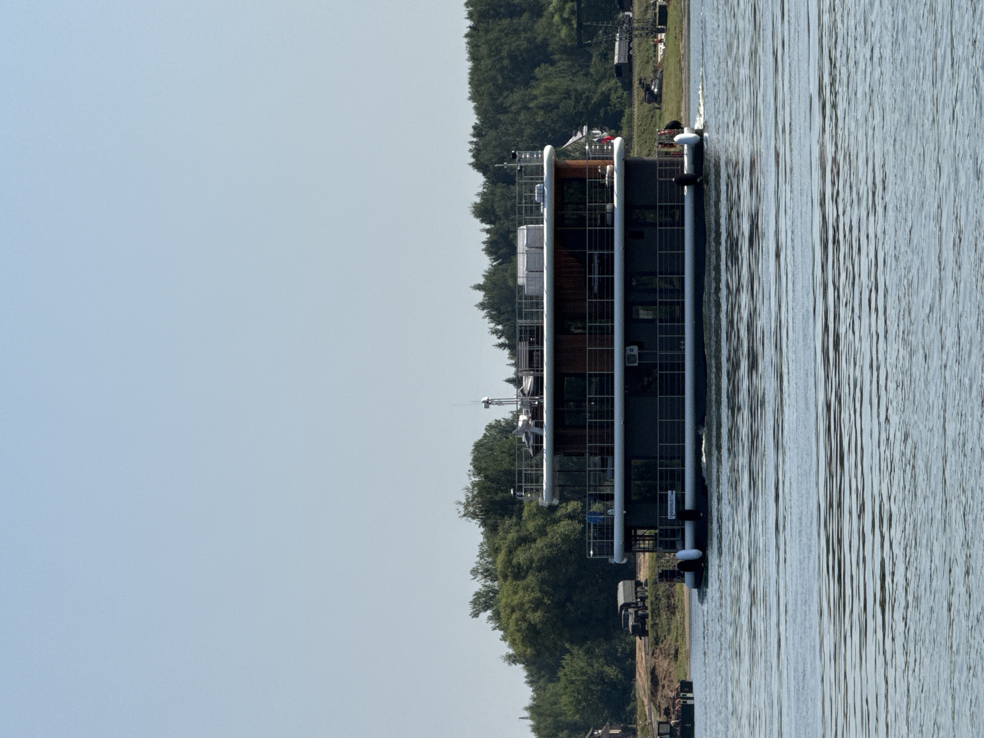 In the city of Rusanovo, Russia, a multi-story house boat is navigating a body of water. The vessel has multiple decks with railings and large windows. On the shore behind the boat, a line of trees forms a dense forest. A dirt road runs along the shoreline, with several military trucks parked on it. The sky is clear and blue. The water is rippling, indicating movement of the boat.
