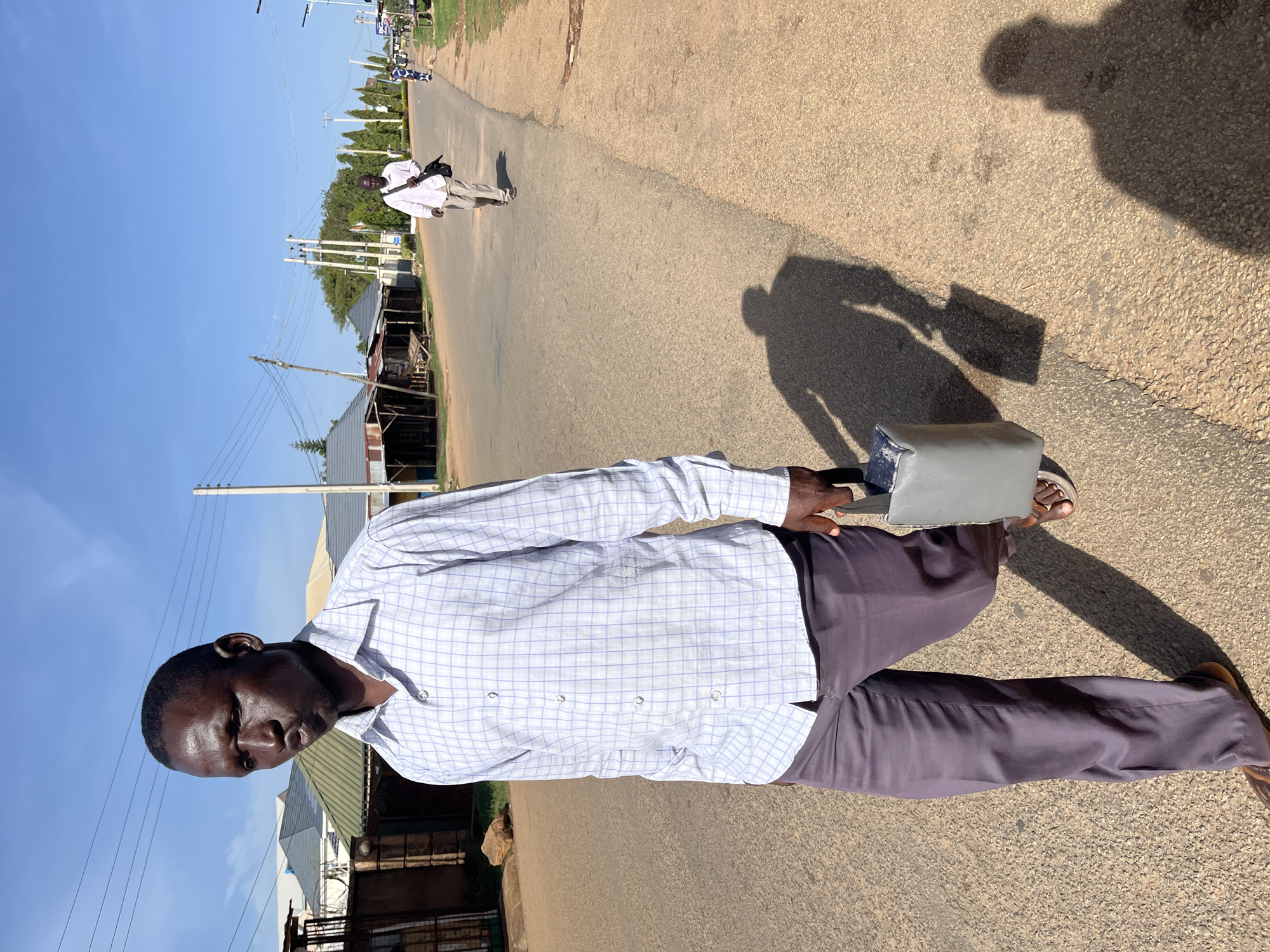 The image depicts an outdoor street scene in Barakin Akawo, Nigeria, under a clear, bright blue sky. The main subject, a man with dark skin and short hair, stands prominently on the right side of the frame, looking directly at the viewer with a neutral expression. He is wearing a light-colored, long-sleeved checkered shirt (white with blue grid lines) and dark grey trousers, paired with open-toed sandals. He holds a grey rectangular bag or satchel in his left hand.
The setting is a street or road with an uneven, granular surface, suggesting a mix of dirt, gravel, and rough asphalt. It extends into the distance, flanked by various buildings, many featuring corrugated iron roofs, indicative of local architecture. Utility poles with numerous overhead power lines are visible along the street, alongside some scattered trees and vegetation.
In the mid-ground, further down the street, another man wearing a light shirt and dark trousers is seen walking away from the camera, carrying a shoulder bag. Other distant figures and structures are also present, contributing to a sense of daily life.
The time of day appears to be midday or early afternoon, characterized by strong, direct sunlight. This is evident from the distinct, elongated shadows cast on the ground, including the shadow of the photographer and the main subject. The lighting creates a high-contrast scene. No visible text is discernible in the image.