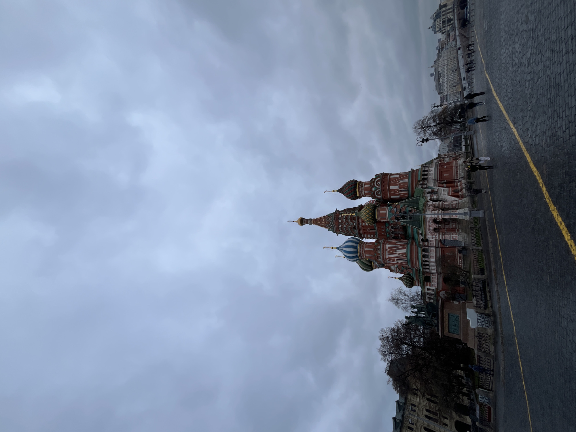 The image shows Saint Basil's Cathedral in Moscow, Russia. The cathedral's colorful domes are prominent in the view. The ground is paved, and a yellow line is visible. A few people are walking along the pavement. Buildings are visible in the background. The sky is overcast.