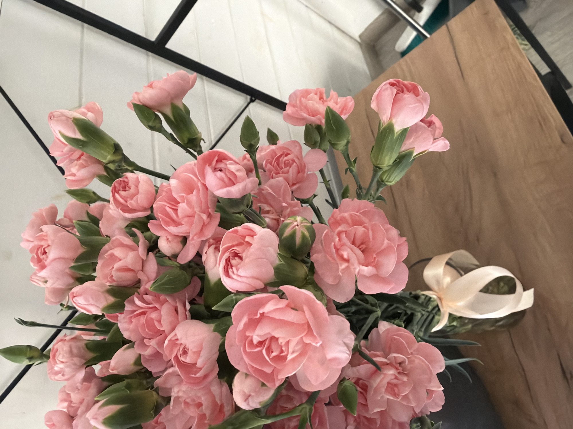 A close-up, high-angle shot shows a bouquet of pink carnations in a clear glass vase on a wooden table. A light-colored ribbon is tied around the neck of the vase. The background is a white, horizontal-paneled wall, with a black geometric frame partially visible on the right. The scene is indoors, likely in a home or studio in Novosaratovka, Russia. There are no people present in the image.