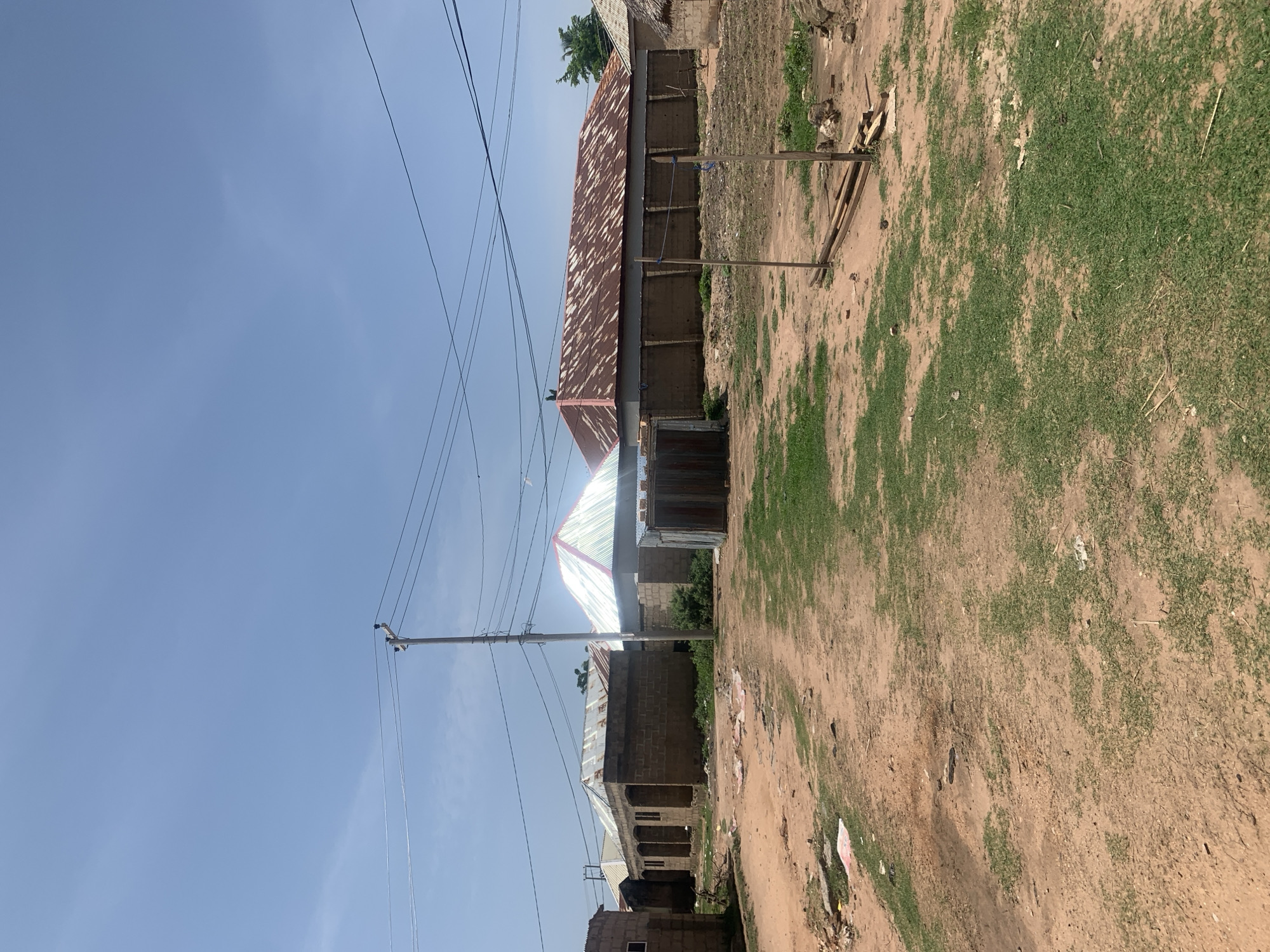 This is a photograph of a street scene in Jauro Yinu, Nigeria. The image depicts several buildings with corrugated metal roofs, constructed from cinder blocks. A dirt road with patches of grass runs through the foreground. In the middle ground, there is a power pole with overhead electrical wires. To the right, there is a wooden structure with what appears to be lumber. The sky is clear and blue.