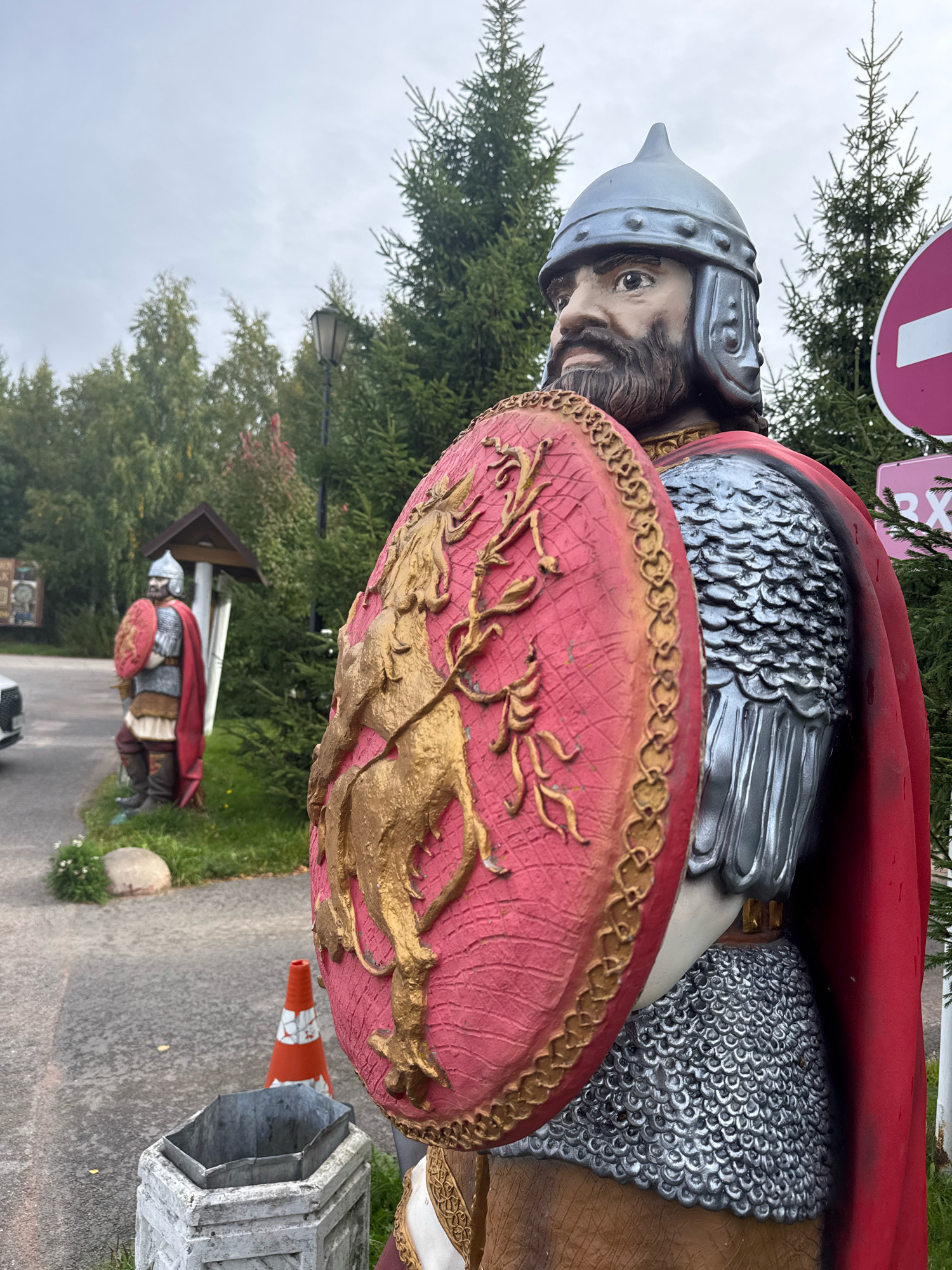 The image shows two life-sized statues of knights in full armor, holding shields and wearing helmets and cloaks. The knight in the foreground is closer to the camera and is positioned on the right side of the frame, with its face looking towards the left. This knight is wearing a silver helmet with a pointed crest, a red cloak, and chainmail armor. It holds a large red shield with a gold emblem of a lion. A second knight, further in the background and on the left side of the frame, is also depicted in armor and holding a shield. The statues are located outdoors, on a paved area with greenery and trees in the background. An orange traffic cone and a metal trash receptacle are visible near the foreground statue. A signpost with a red and white circular symbol is partially visible on the right side of the frame. The sky is overcast. The general atmosphere suggests an outdoor display or attraction, possibly related to historical reenactments or themed decoration in a public space. The specific location is identified as Peterhof, Russia, implying a possible connection to the historical or cultural attractions of that area.