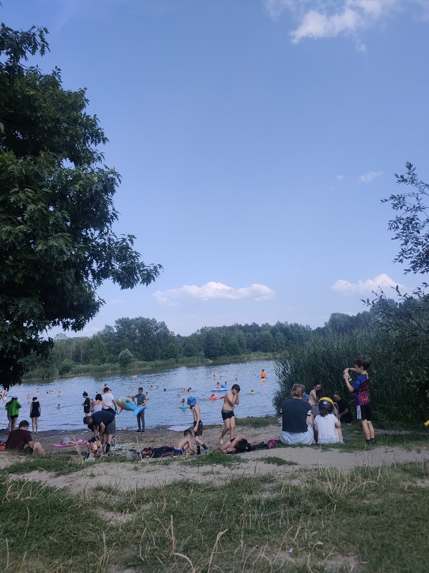The image depicts a bustling scene at a lake, likely a popular recreational spot in Kobyłka, Poland, on a clear, sunny day. In the foreground, a sandy embankment is dotted with people relaxing and preparing for water activities. Some are seated on towels, while others are engaged in conversation or tending to their belongings. A group of individuals are gathered near the water's edge, some wading into the lake.
The lake itself is populated with swimmers and individuals engaged in various water sports. In the middle ground, a man is carrying a large inflatable dolphin, suggesting a playful atmosphere. People are spread across the water, some with inflatable rings and devices. The background is characterized by a dense line of trees and vegetation bordering the lake, typical of the natural landscape surrounding Kobyłka. The sky is a bright blue with scattered white clouds, indicating fair weather. The overall impression is one of a lively summer day at a public lake.