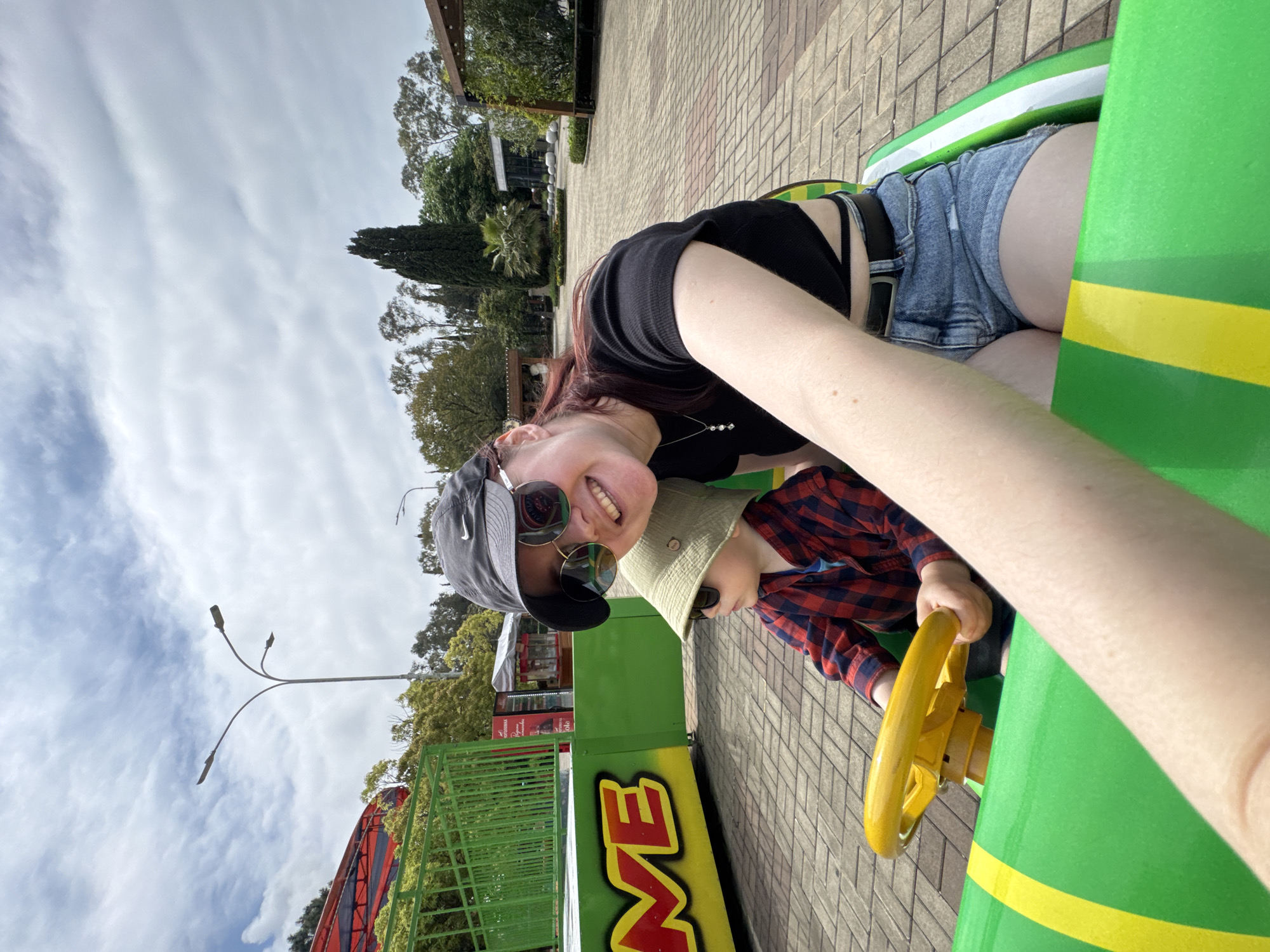 A medium, off-kilter shot captures a woman and a child riding an amusement park ride. The woman is on the left, sporting sunglasses, a black top, and denim shorts. She's smiling at the camera. To her right is a child wearing a hat and a red plaid shirt, gripping the steering wheel of the ride. The ride is green with a yellow stripe. The backdrop reveals brick pavement, trees, streetlights, and buildings. Based on architectural elements and context, it's highly probable that the location is in the city of Sochi, Russia.