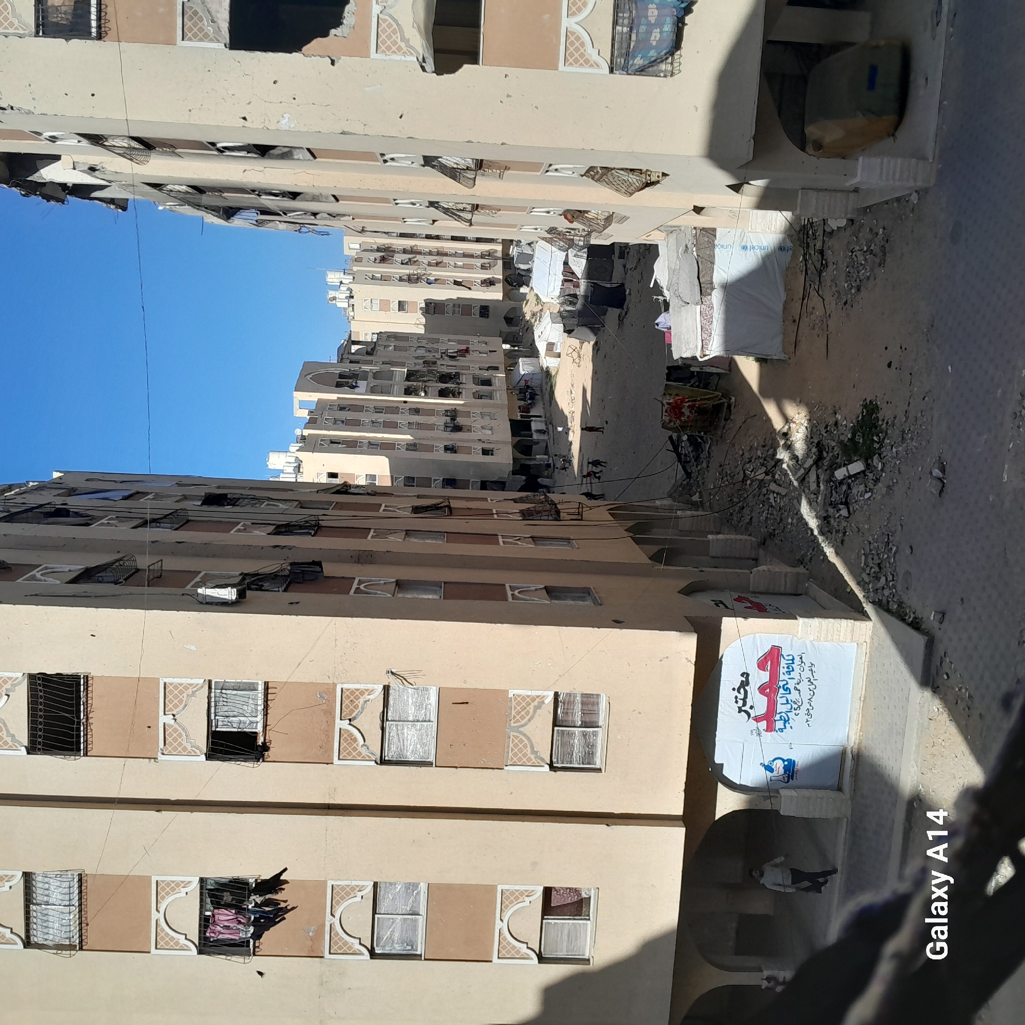 The image depicts a narrow street in a city, possibly in the Middle East, lined by tall, weathered apartment buildings. The buildings have multiple stories with windows that feature decorative patterns. Damage to the buildings is evident, with broken windows and signs of decay. Clothes are hung outside some windows, suggesting residential use.
In the street, there appear to be makeshift shelters or tents, indicating a possible refugee camp or displacement situation. Debris and rubble are scattered on the ground, further suggesting damage or conflict.
There is a sign or banner with Arabic script visible in the foreground, possibly advertising medical services. A person is seen walking near the sign. The sky is clear and blue. The image appears to have been taken with a "Galaxy A14" smartphone.