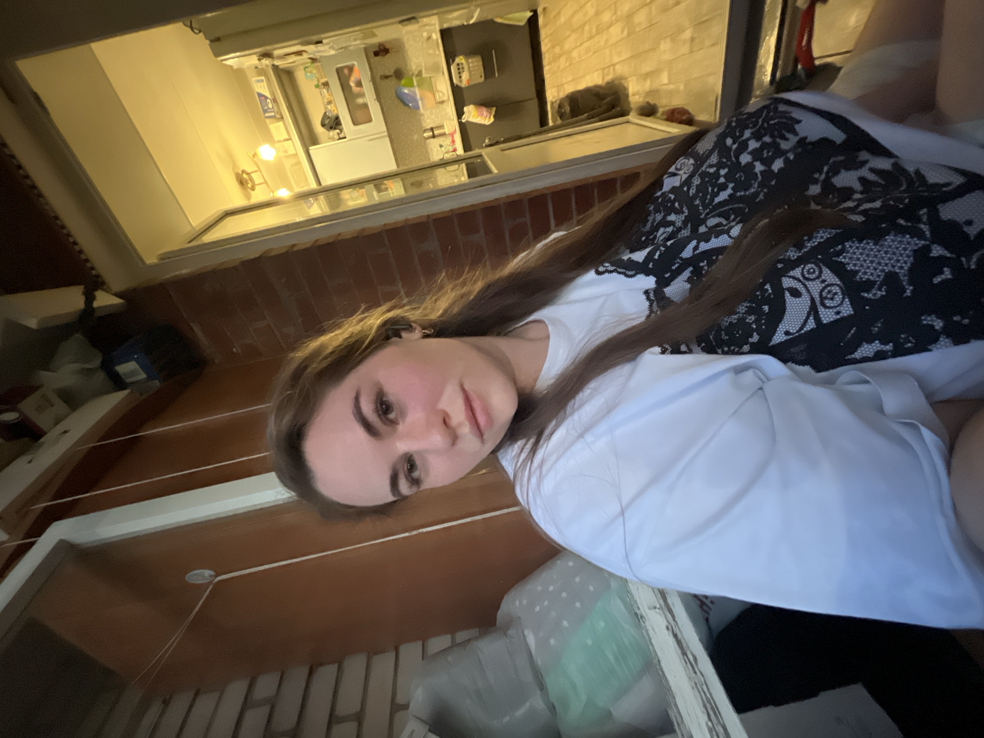 The image shows a woman with long brown hair lying in what appears to be a bed or resting area. She is wearing a white top with a black lace overlay. Behind her, a window provides a view of a kitchen area. The kitchen contains white cabinets, a refrigerator, and other kitchen appliances. The exterior of the building appears to be brick. The specific location is Chaikovsky, Russia.