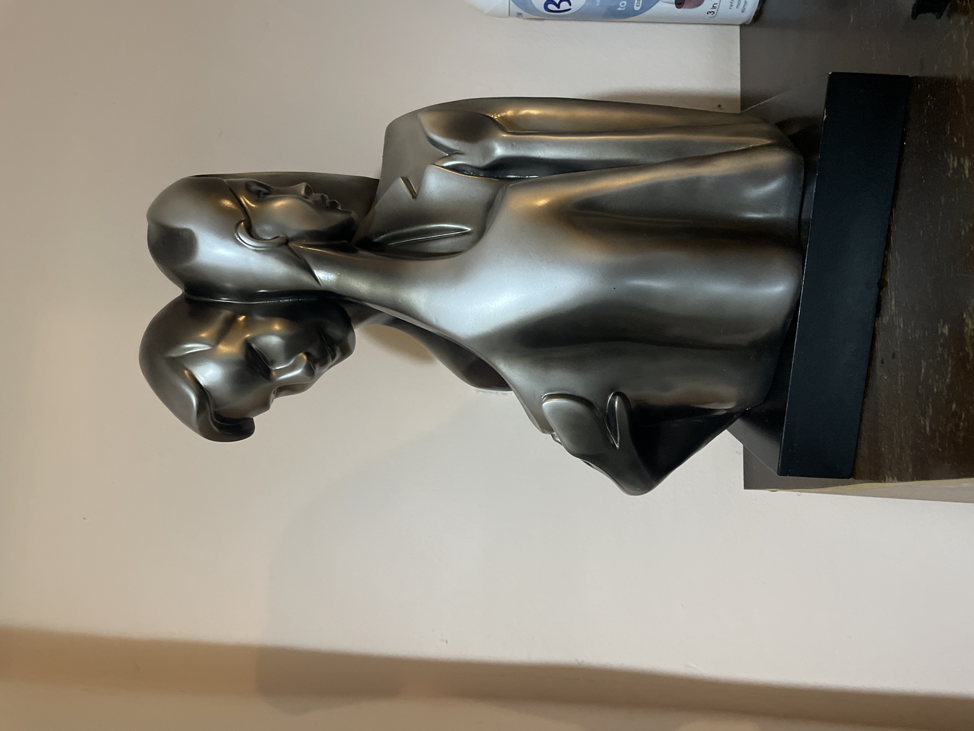 A close-up, indoor shot shows a silver-toned sculpture of two human figures intertwined on a dark, rectangular base. The background is a plain, off-white wall. At the top right, a white and blue bottle is partially visible. No information about Yerevan, Armenia is discernable.