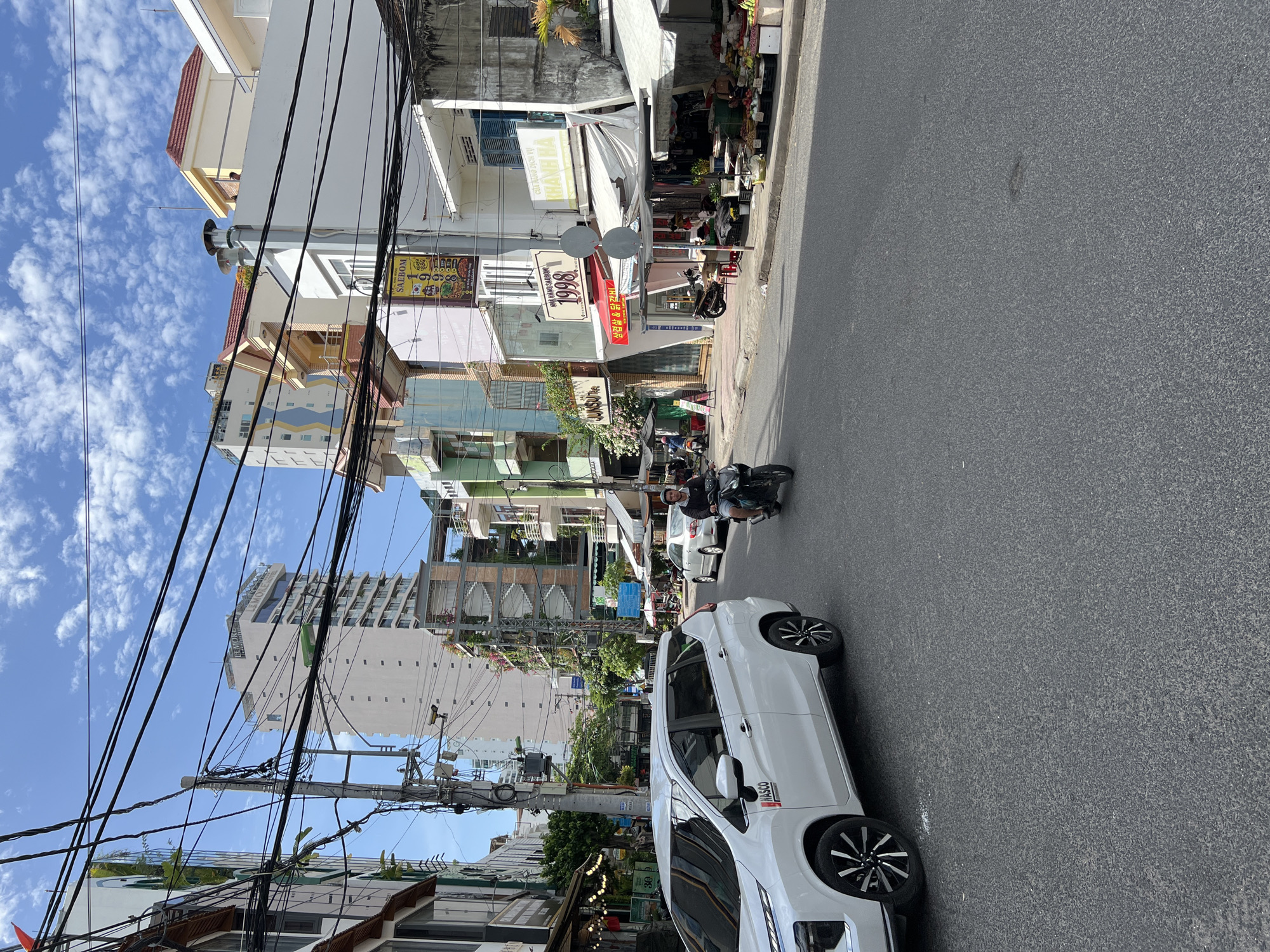 The image depicts a street scene in Nha Trang, Vietnam, characterized by a mix of modern and older buildings, dense overhead power lines, and vehicular traffic. A white SUV is parked on the left side of the road. A man is riding a motorcycle in the right lane, heading away from the camera. The street is lined with various establishments, including shops and restaurants, some with awnings and displays of goods. A fruit stand is visible on the right sidewalk. The sky is blue with scattered white clouds. The buildings vary in height and architectural style, with some appearing to be residential and others commercial. The overall impression is of a bustling urban environment.