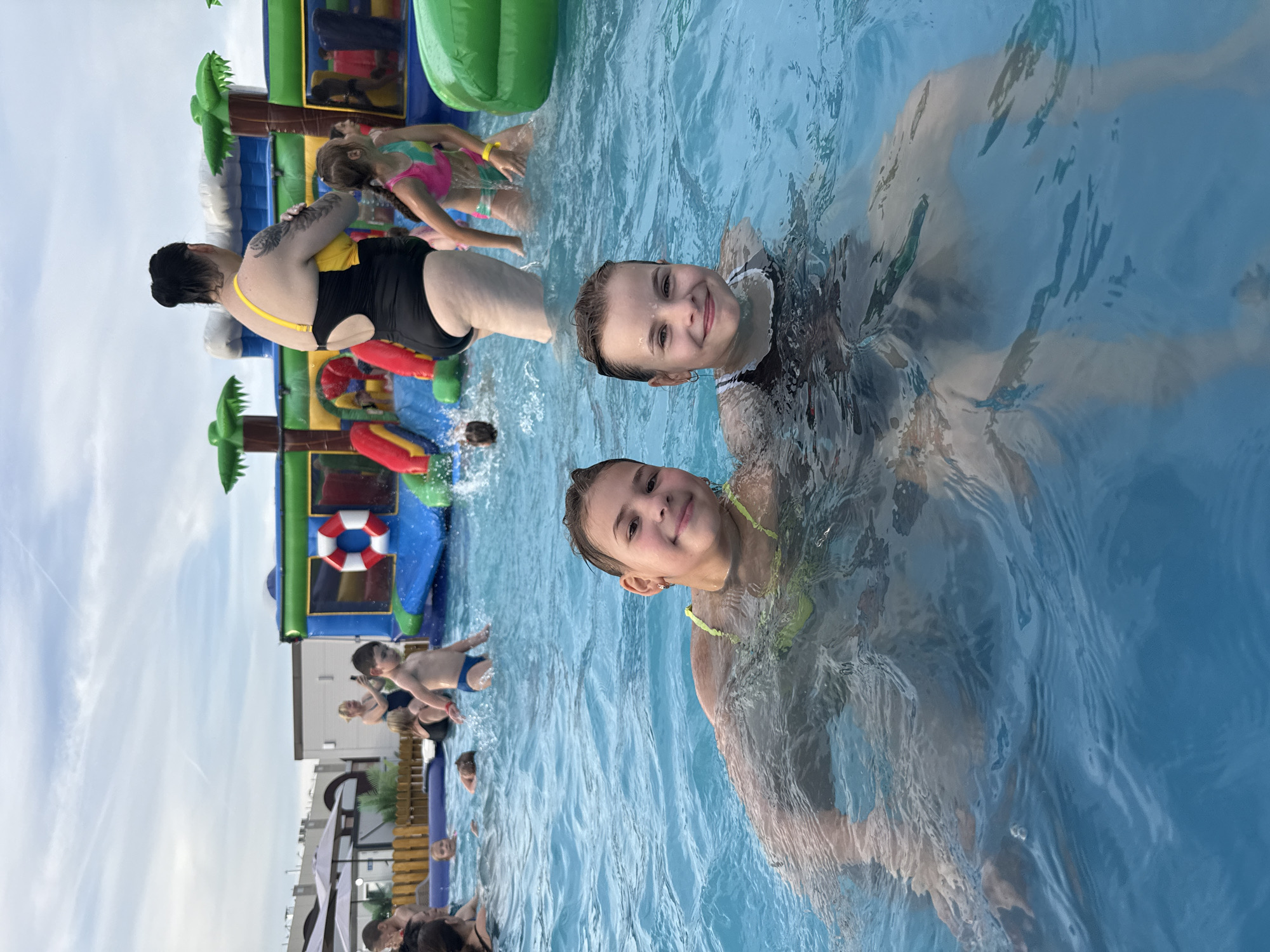 In a bright outdoor swimming pool, several people are enjoying a day of recreation. Two young girls are the primary subjects, captured as they swim. One girl, with a bright smile and light brown hair, is looking directly at the camera, her face partially submerged in the water. She is wearing a yellow swimsuit. The other girl, also with light brown hair and a similar smile, is positioned slightly behind and to the left, also in the water.
In the background, a large inflatable water park with vibrant colors and palm tree decorations is set up in the pool. Several other individuals, including children and adults, are visible enjoying the water and the inflatable structures. One woman, with dark hair pulled back and wearing a black and yellow swimsuit, is partially submerged near the inflatable. A young boy in blue swim trunks is also visible. The water is clear and blue, with ripples indicating movement.
The scene suggests a sunny day, likely during daylight hours, conducive to outdoor swimming activities. The presence of the inflatable water park indicates a fun, recreational event, possibly a party or a family outing. The overall mood appears to be joyful and relaxed, with people of various ages engaging in water-based activities. The setting is an outdoor pool, possibly at a resort, water park, or a large residential complex, with a clear sky visible in the background. There is no visible text in the image.