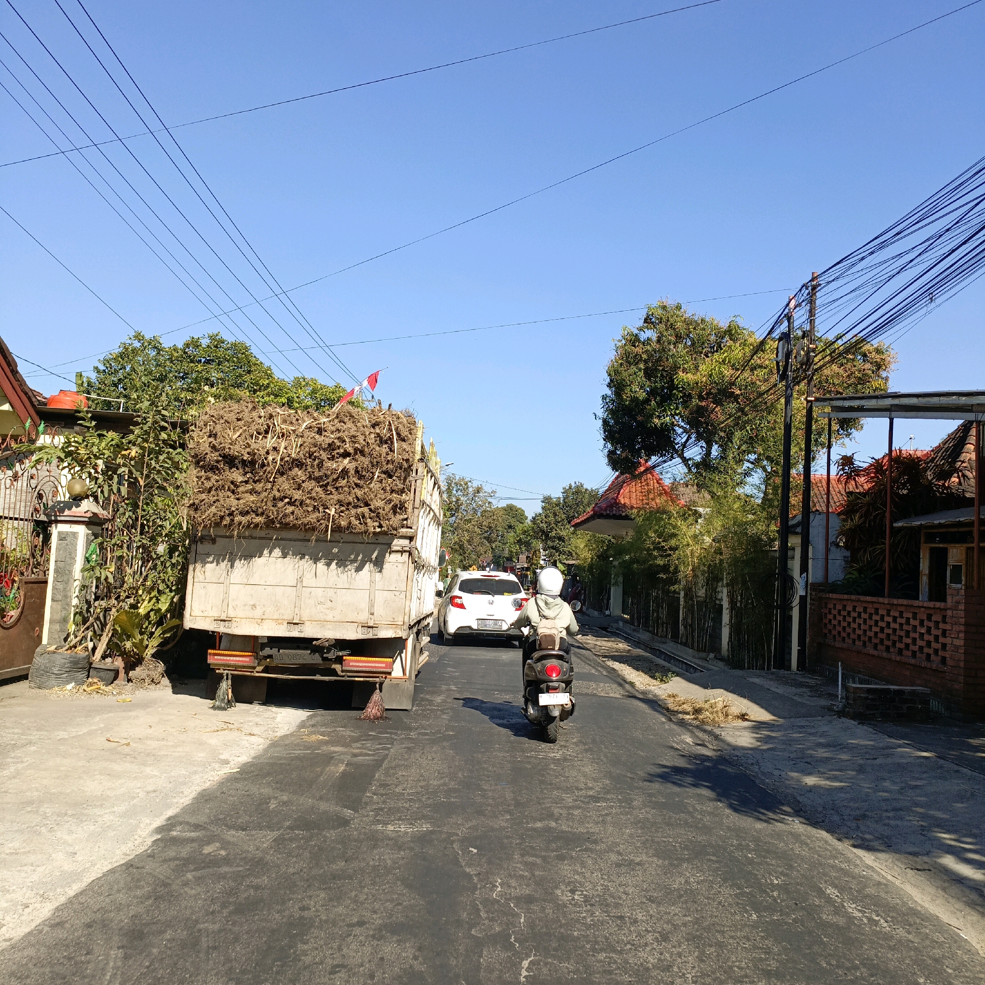 The image captures a street scene in Sraten, Indonesia, under a clear blue sky. A large truck laden with dry roots or plant matter is parked on the left side of the road. A white car is moving forward in the same lane as the truck. A person on a scooter, wearing a white helmet and backpack, is riding in the lane behind the white car, also moving forward. The road appears to be paved with asphalt, with visible repairs or patching. The sides of the road feature concrete sidewalks and some vegetation. Buildings with red-tiled roofs and brick walls are visible along the right side of the street, interspersed with trees and bamboo. Power lines are strung across the sky, supported by utility poles. The overall impression is of a typical day on a moderately busy street in a semi-urban or rural Indonesian setting.