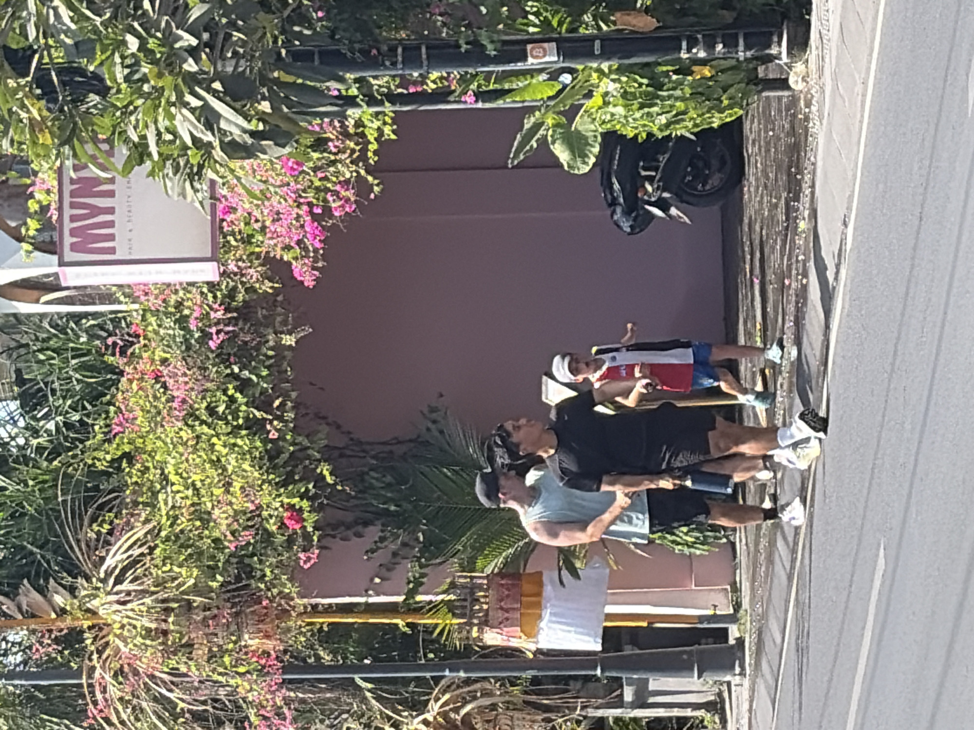 A group of three people, two men and one child, are walking on a sidewalk next to a street. The man in the front is wearing a black t-shirt and black shorts. The man behind him is wearing a light blue tank top and black shorts. The child, walking slightly ahead of the men, is wearing a white shirt with red and blue trim and blue shorts. They are walking in front of a pink building with a sign that reads "MYN HAIR & BEAUTY SPA." Lush green plants and pink flowers are growing on the building and around the sidewalk. A dark-colored motorcycle is parked on the sidewalk behind the people. The ground on the sidewalk appears wet and reflective, possibly from recent rain or cleaning. The scene is illuminated by bright sunlight, casting shadows on the ground. The location is North Kuta, Indonesia.