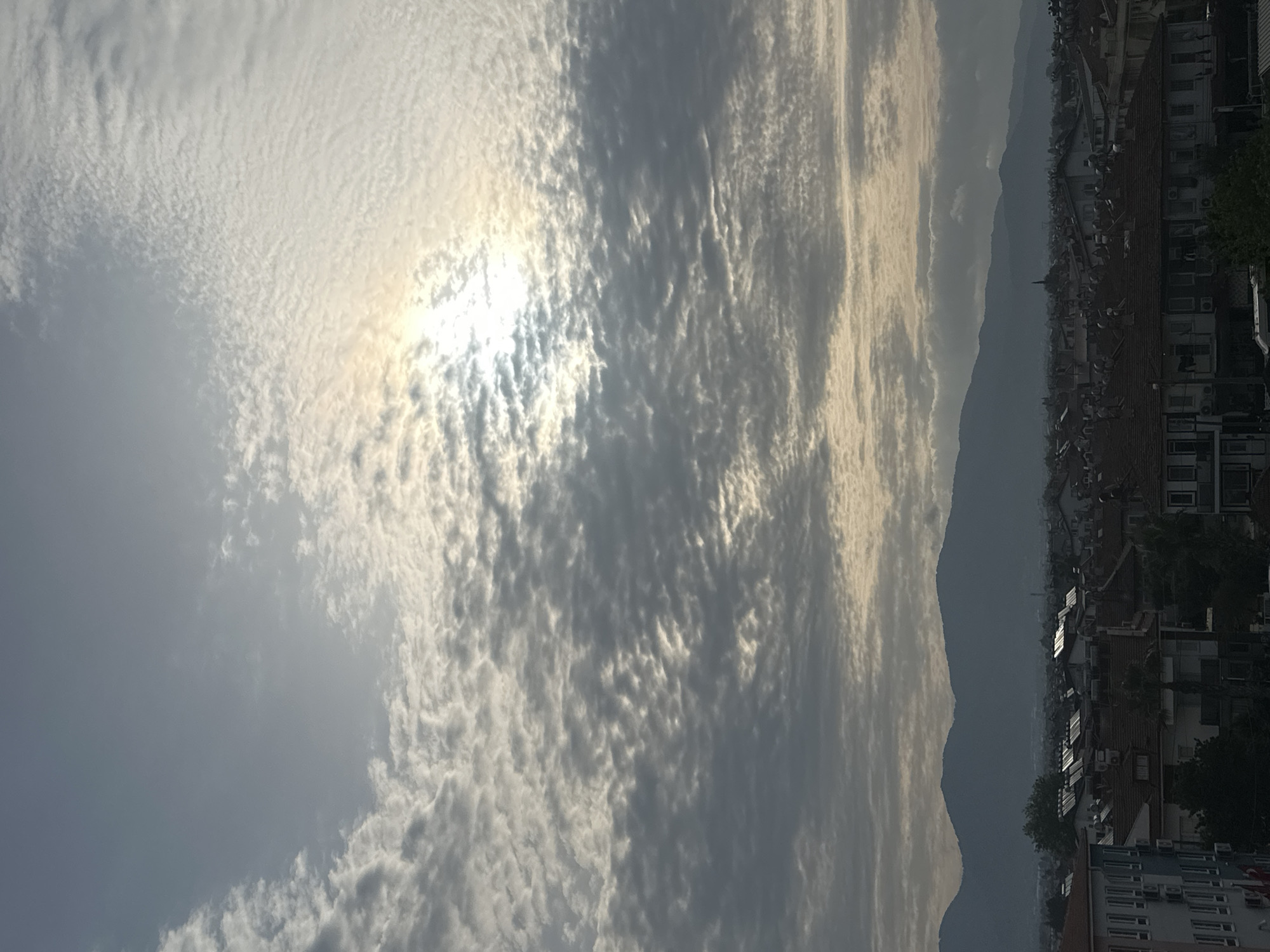 The image captures an urban view in Fethiye, Turkey, under a sky filled with altocumulus clouds partially obscuring the sun. Mountains form a backdrop to the city skyline. The urban scene features rooftops, buildings, and trees. There are no people visible.