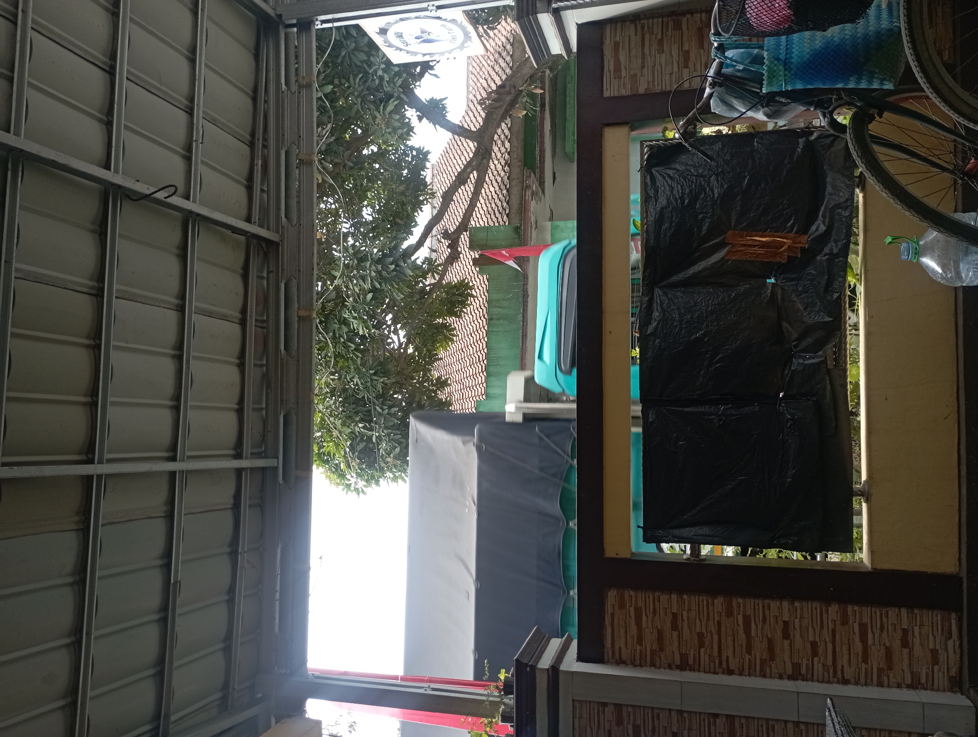 The image depicts an outdoor scene, likely from a porch or covered outdoor area, looking out towards a street. In the foreground, there is a bicycle with two colorful bags attached to the handlebars, one blue and white checkered, and another more loosely hung. A large plastic water bottle is on the ground near the bicycle. A black plastic tarp is stretched across a fence or barrier, with a piece of orange material hanging from it. Beyond the fence, a large truck with a dark tarp covering its cargo is visible, as well as a turquoise-colored object. Trees with green leaves and a tiled roof of a building are also present. A sign with blue text and a logo is visible on the right side of the frame. The overall setting suggests a residential or commercial area in Maos Kidul, Indonesia.