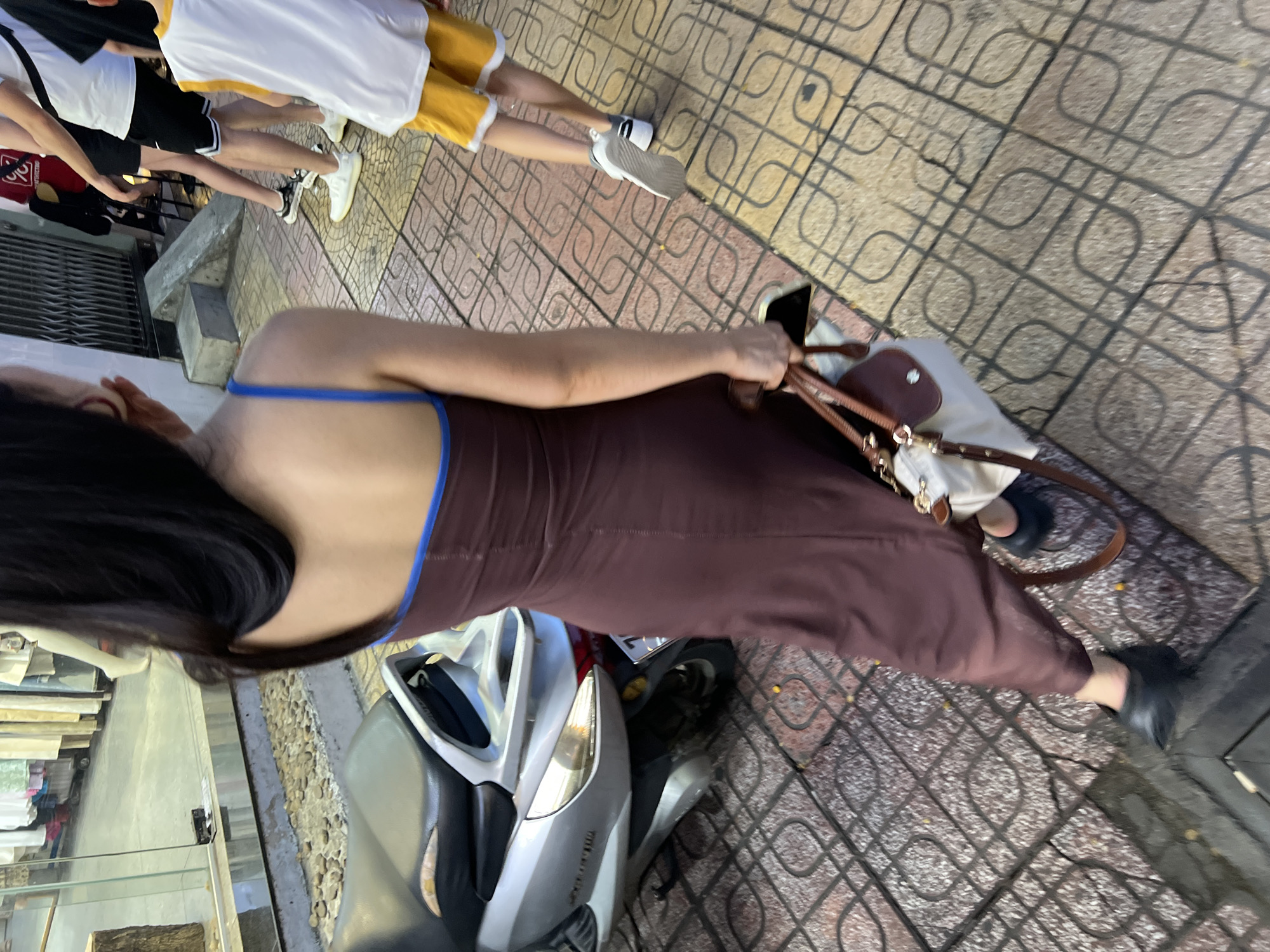 A medium shot, taken from a high angle, captures a woman in a dark brown, floor-length, strappy dress with a blue trim on the neckline. She is walking on a tiled sidewalk in Nha Trang, Vietnam. The woman is carrying a light-colored tote bag with brown leather accents and a smartphone. To her left, a silver scooter is parked. In the background, a shop window displays clothing on mannequins. Other pedestrians are visible further down the street. The sidewalk is made of patterned concrete tiles.