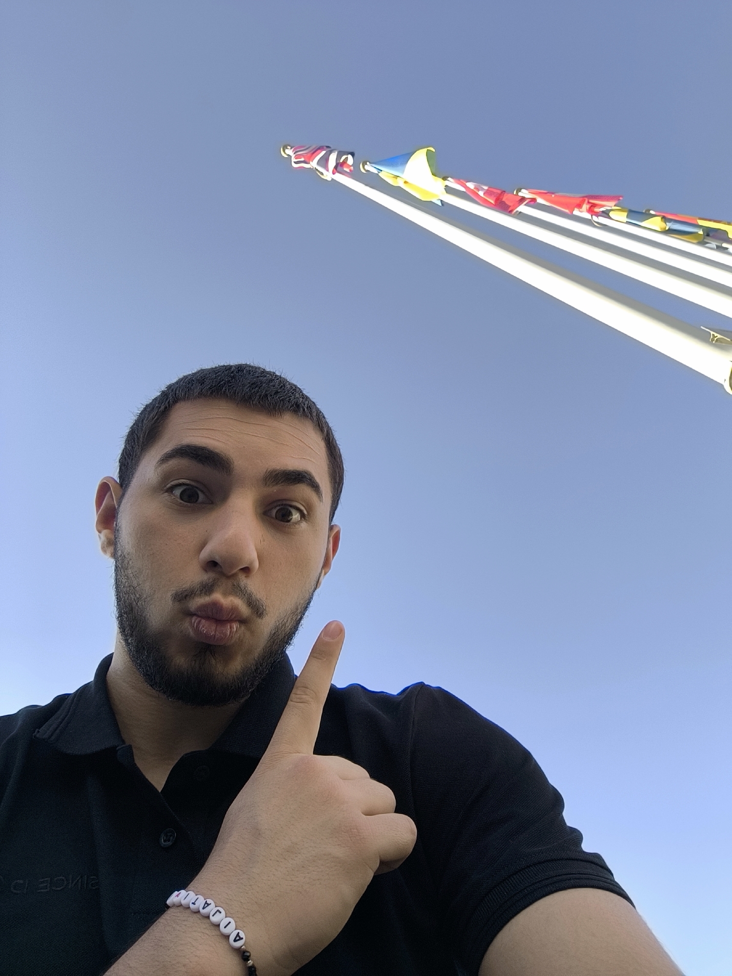 A young man is featured in a close-up, low-angle selfie, positioned in the lower-left corner of the frame. He has dark hair and a beard, and his lips are pursed in a playful pout. He points upwards with his right index finger. He is wearing a black collared polo shirt, with inverted text "SINCE D" visible on the fabric near his left shoulder. On his right wrist, he wears a white beaded bracelet with black letters that spell "A JOAN".
The background is dominated by a clear, bright blue sky. Diagonally across the upper right, multiple national flags are seen flying from tall, white flagpoles. While not all flags are fully discernible, visible colors include red, white, blue, and yellow, with one appearing to be the Union Jack (United Kingdom) and another a blue and yellow flag (possibly Ukraine). The presence of several national flags on flagpoles in Strasbourg, France, strongly suggests a location related to international European institutions like the Council of Europe or the European Parliament. The clear sky indicates daytime.