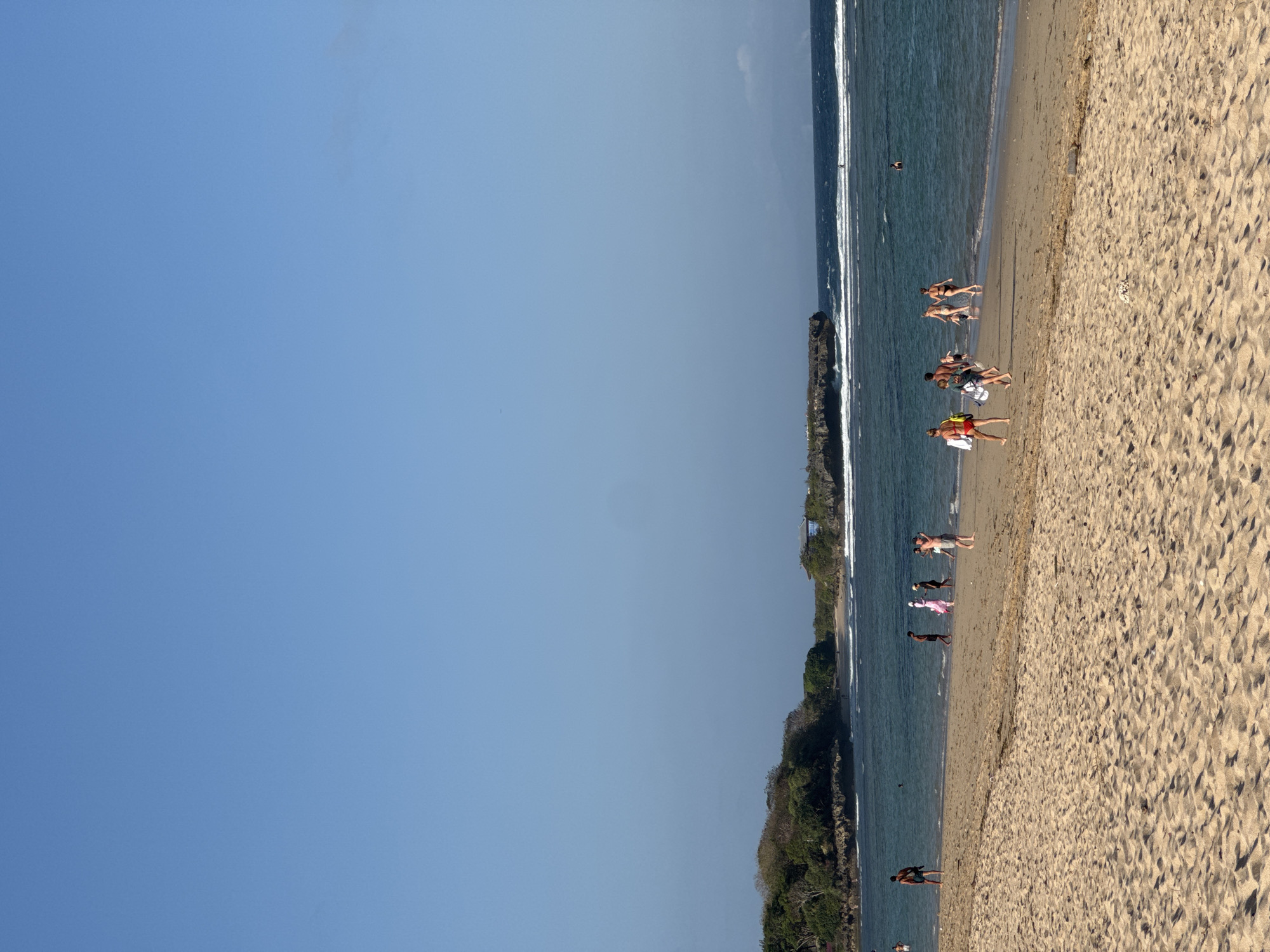 The image depicts a wide, sandy beach in Kuta Selatan, Indonesia, with a clear blue sky overhead and the ocean in the background. Several individuals are visible on the beach, some walking along the shoreline and others wading in the shallow water. The beach has gentle waves rolling in, and the water appears a deep blue. On the left side of the image, a lush green hillside with trees slopes down towards the water. Further in the distance, a rocky outcropping rises from the sea. The scene is bathed in natural sunlight, suggesting it is daytime.