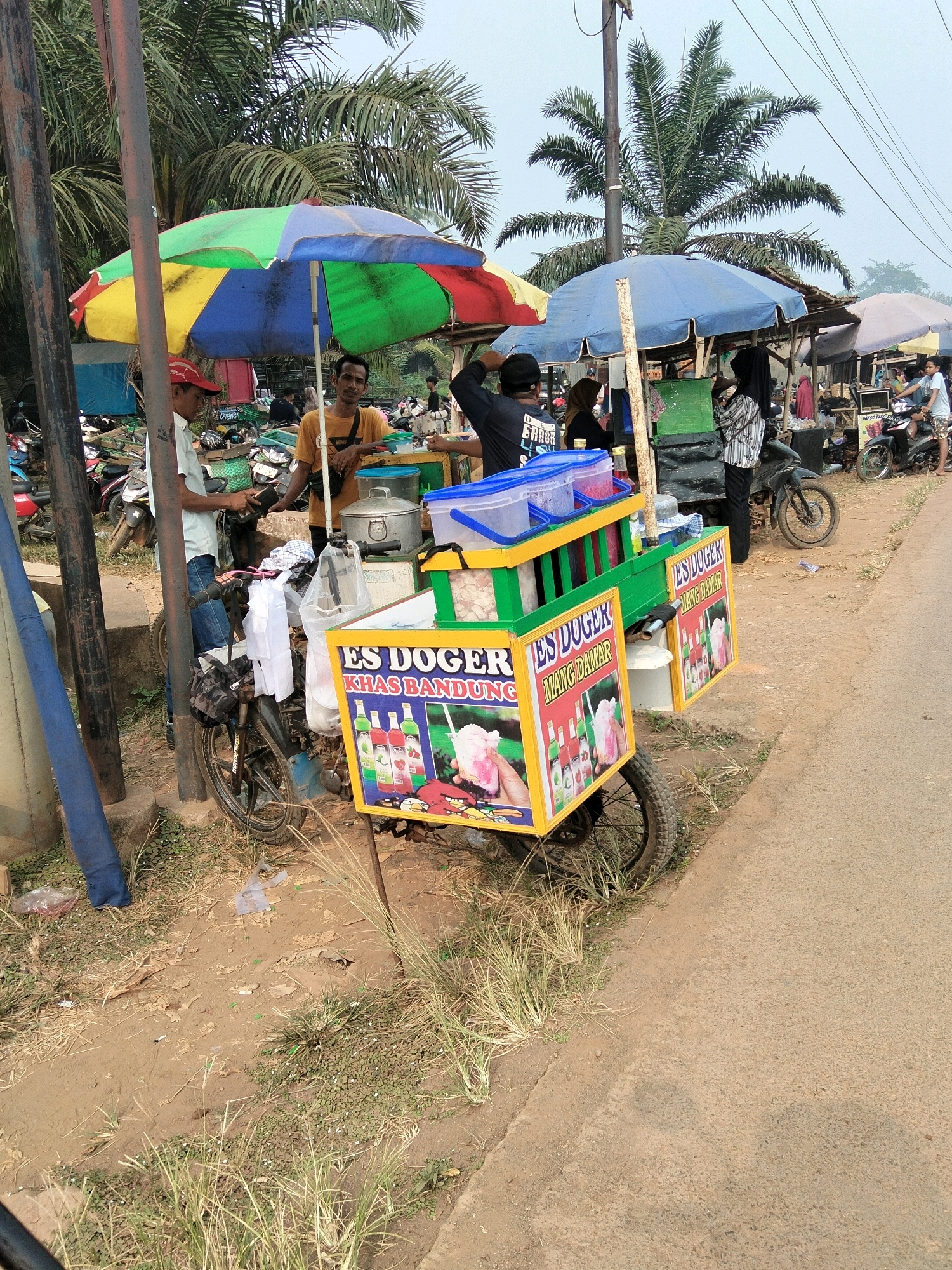 The image depicts a roadside scene in Tirto Sari, Indonesia, likely a marketplace or a bustling street corner, given the presence of several food stalls and parked motorcycles. In the foreground, a colorful, multi-paneled umbrella shades a street food cart selling "Es Doger". The cart itself is decorated with vibrant images of the product and text in Indonesian. Two men are visible near the cart, one in an orange shirt interacting with the cart, and another in a light blue shirt standing by a motorcycle. Behind them, other stalls are set up, with more people browsing and interacting. A prominent blue umbrella shades another stall in the mid-ground. The environment appears to be outdoors, with palm trees visible in the background and a paved road running alongside the stalls. The ground is a mix of dirt and dry grass, suggesting a warm, possibly humid climate. Several motorcycles are parked haphazardly, indicating transportation is a common mode for vendors and customers alike. The overall atmosphere suggests a lively, informal commercial setting.