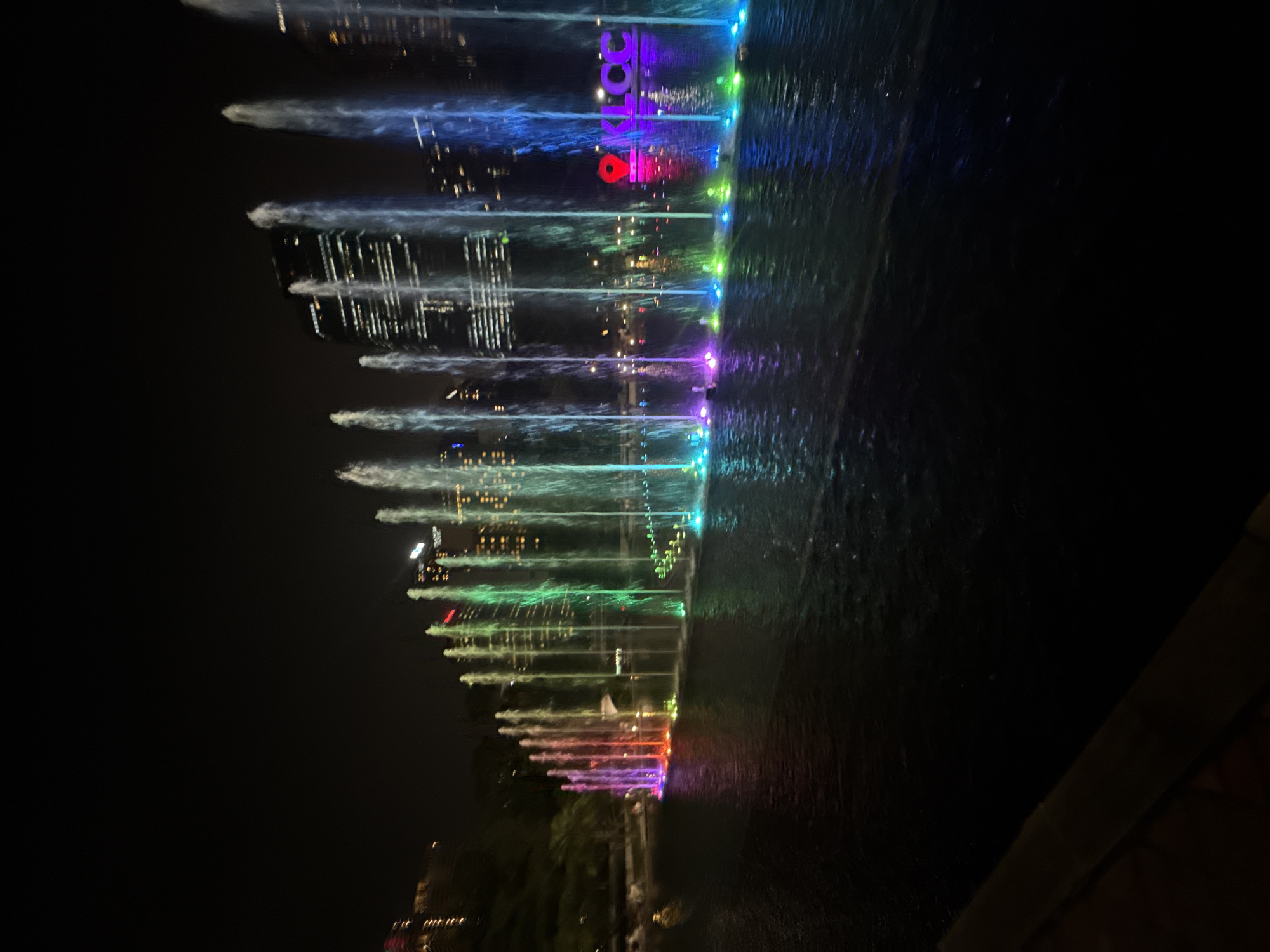 The image displays a vibrant nighttime scene of an outdoor water fountain show in a city setting. The photograph is oriented vertically, showing numerous jets of water shooting upwards from a body of water. Each water jet is illuminated with a dynamic array of colors, transitioning from cool blues and greens on the left to warm reds, oranges, and purples on the right. These colorful lights are vividly reflected on the dark surface of the water, creating a mesmerizing visual effect.
In the background, several tall, modern buildings are visible, their windows glowing with lights against the dark night sky, indicating a bustling urban environment. A prominent feature within or just beyond the fountain display is an illuminated sign spelling "KLCC" in red and purple/pink letters, accompanied by a red location pin icon. This text clearly identifies the location as Kuala Lumpur City Centre.
The scene captures a lively event, likely a choreographed light and water show, typical of a public attraction. The time of day is undeniably night, with artificial lighting providing all the illumination. The sky is dark and clear, and there are no people visible in the frame, allowing the focus to remain entirely on the colorful spectacle of the fountains and the urban backdrop.