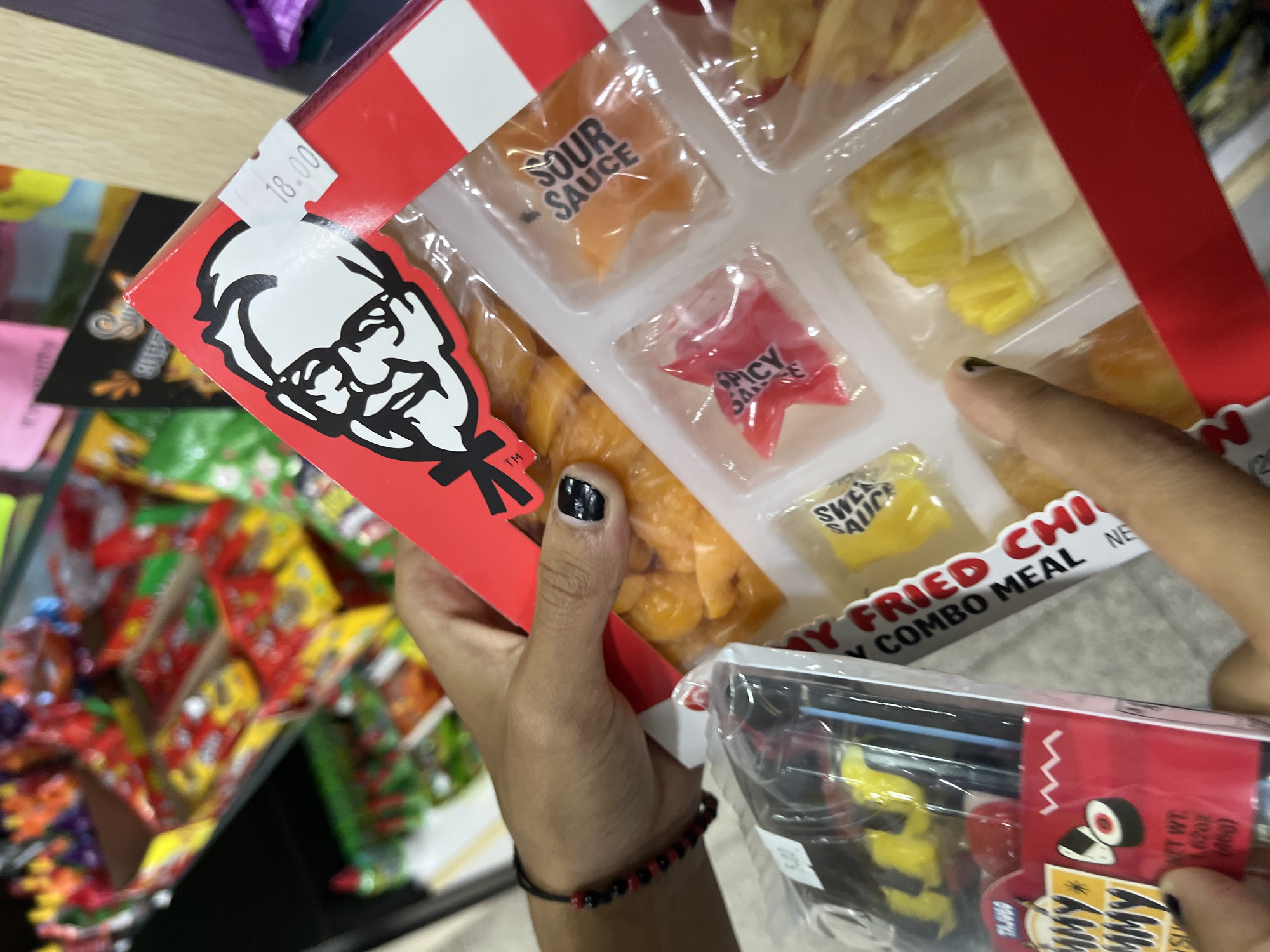 A person holds a KFC-branded candy pack containing gummies shaped like various items from a combo meal. The candy pack features gummies in the shapes of sour sauce, spicy sauce, and sweet sauce in addition to the chicken. The price is marked at 18.00. In the background, other candy products are visible. The person's hand has black nail polish and a beaded bracelet. The brand name "YUMMY" is partially visible on another candy box. The location is likely a store somewhere in Porlamar, Venezuela.
