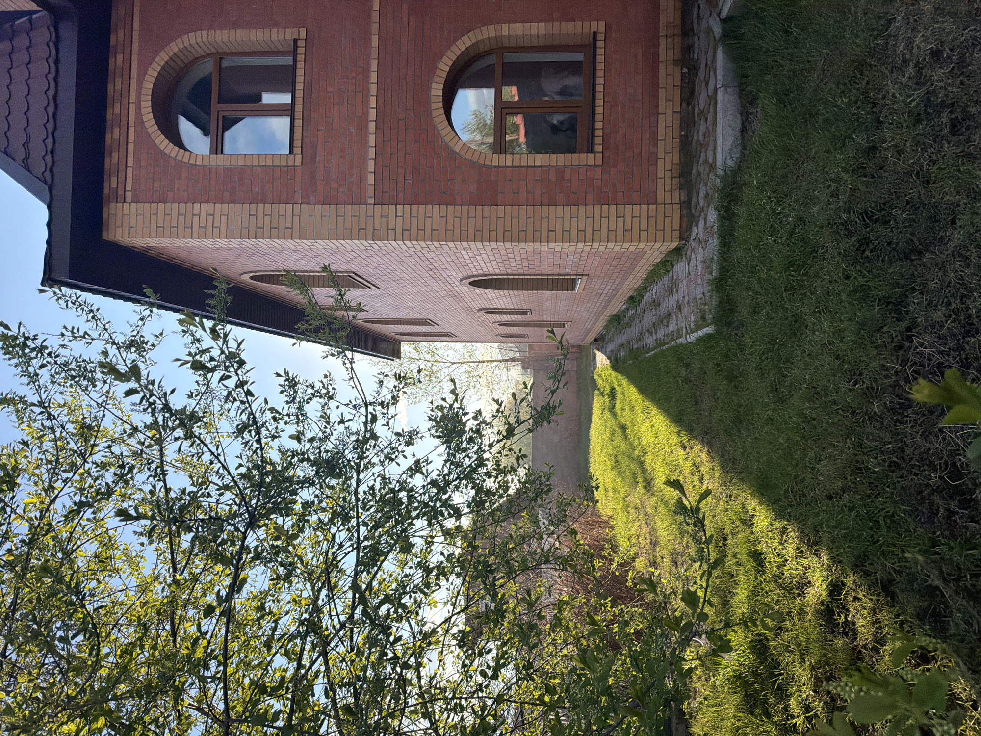 The image shows a building made of red and yellow bricks. The building has arched windows. Next to the building is a grassy area. A tree with green leaves is in the foreground. The location is Krasnoyarsk, Russia.