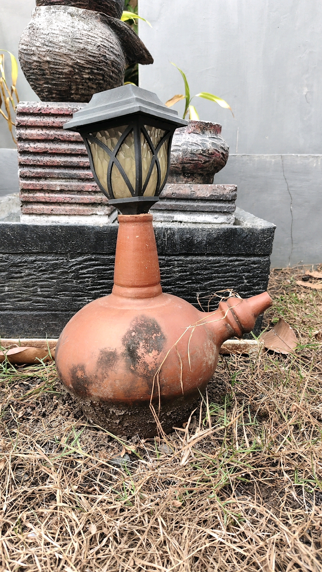 A terra cotta water jug with a lamp on top is placed on the ground in a garden in Banjarmasin, Indonesia. The jug is round and bulbous with a narrow neck that extends upwards to support the lamp. The lamp has a black metal frame and a textured glass shade. The jug is weathered and shows signs of age and use. The ground around the jug is covered in dry grass and leaves, and in the background, there is a wall and some potted plants. The scene appears to be a decorative element in a garden.